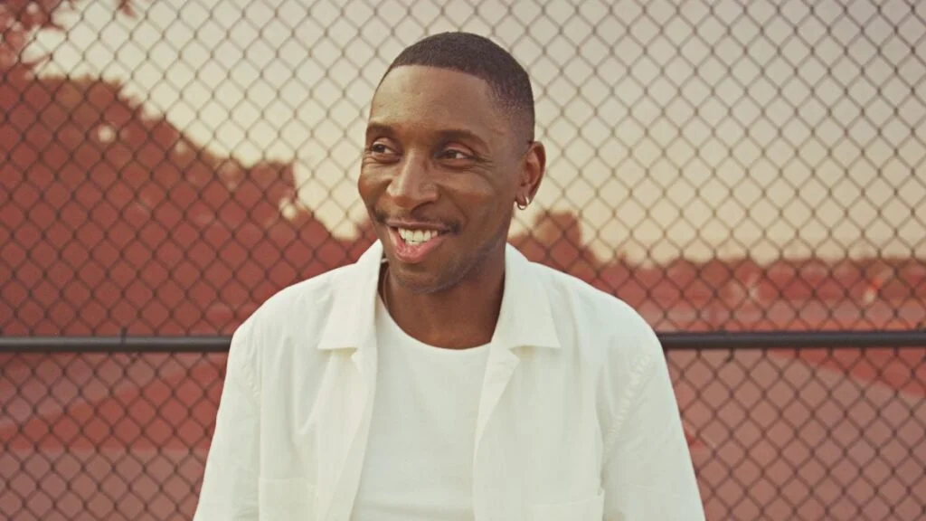A smiling man outdoors standing in front of a chain-link fence with red roof tiles in the background.