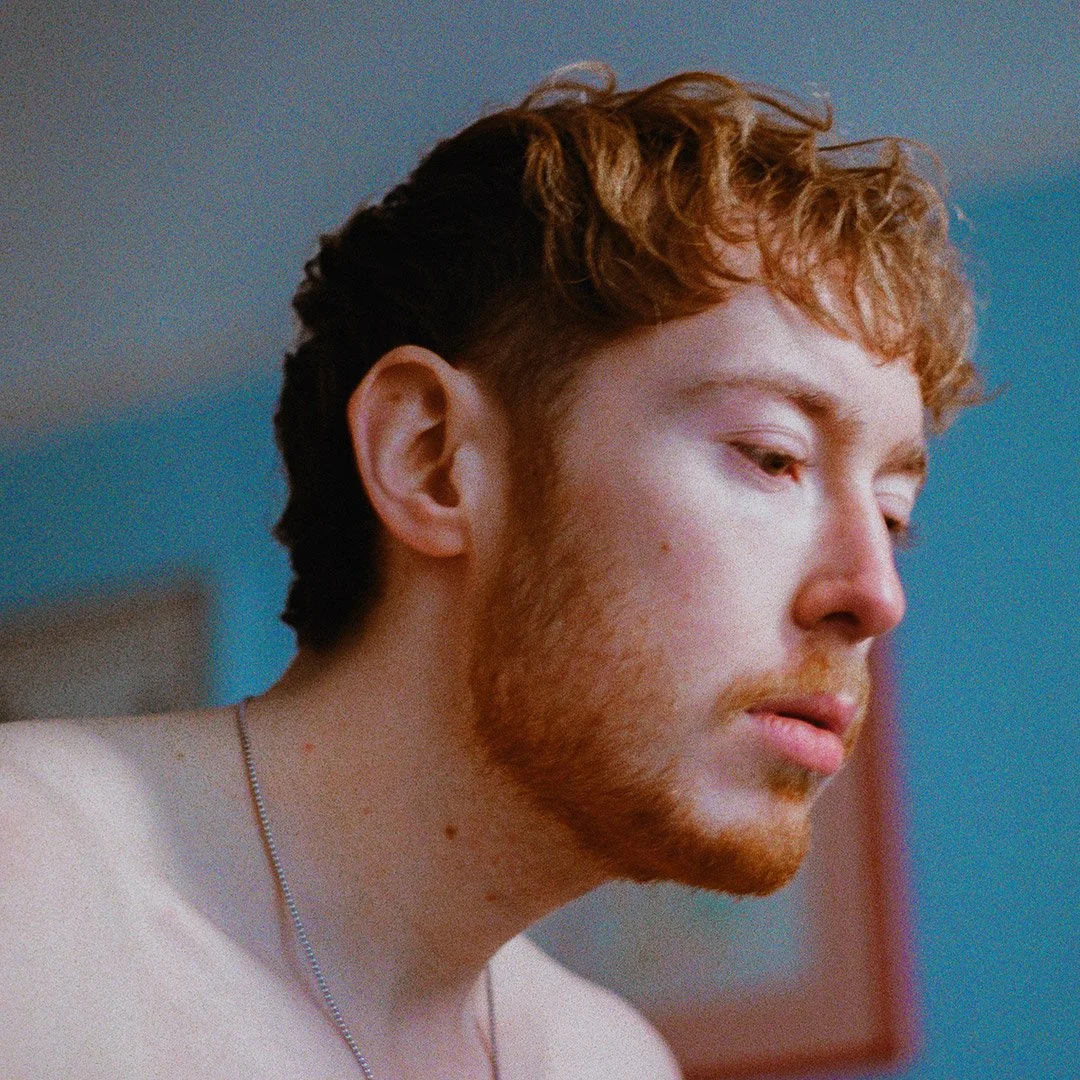A close-up of a young man with red hair and beard, looking downward in a contemplative or somber mood, indoors with a blue wall in the background.