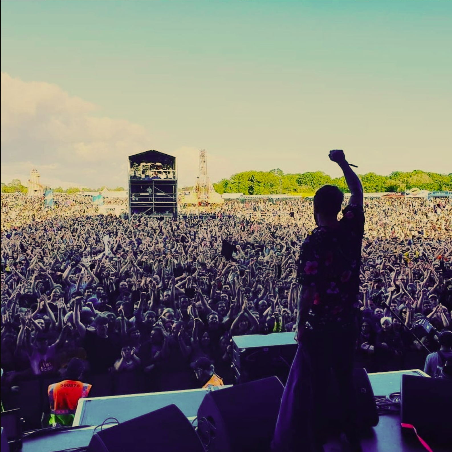 A performer on stage raising their right arm in front of a large audience at an outdoor concert, with festival rides and attractions visible in the background on a sunny day.
