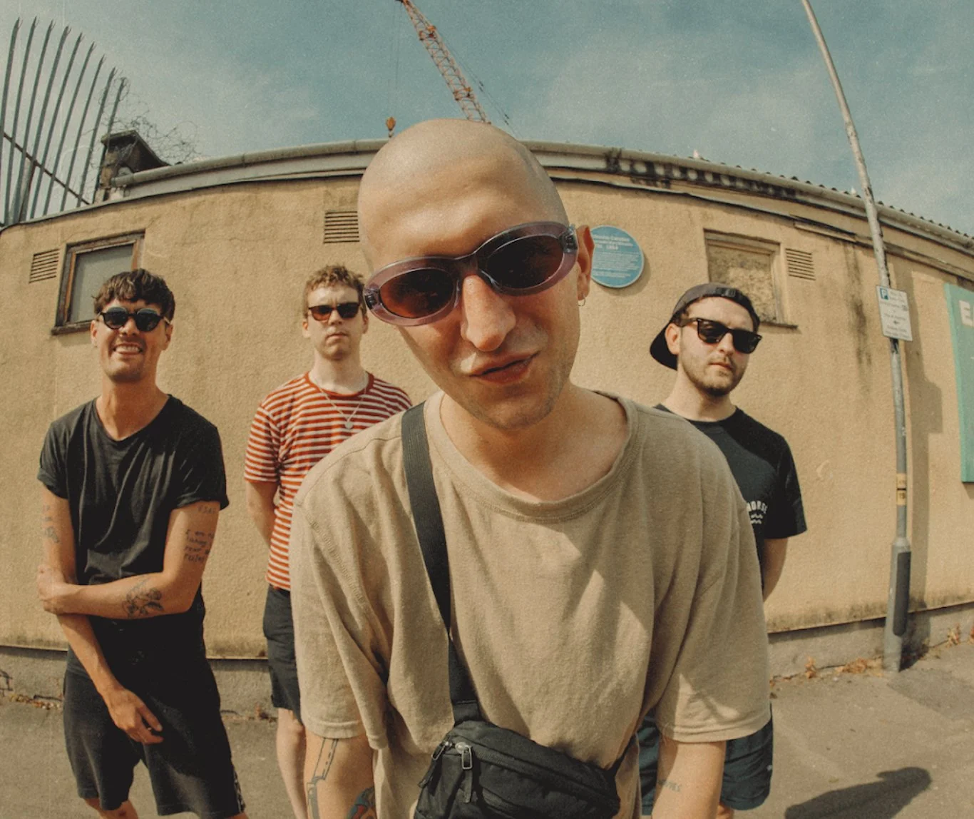 Four young men standing outdoors against a beige wall, wearing sunglasses and casual clothes on a sunny day, with a construction crane in the background.