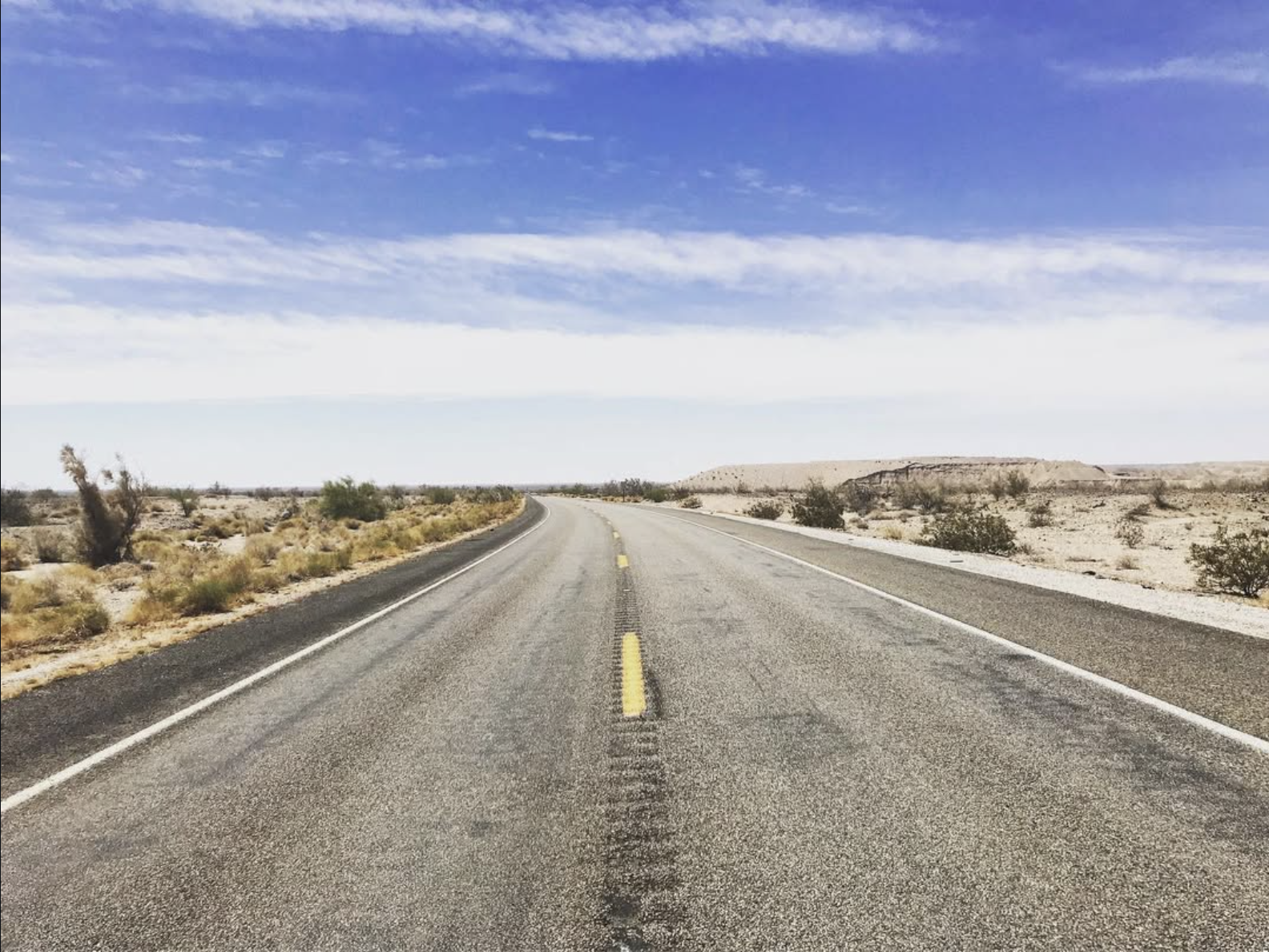 Empty desert highway stretching into the horizon under a partly cloudy blue sky with sparse desert vegetation on both sides.
