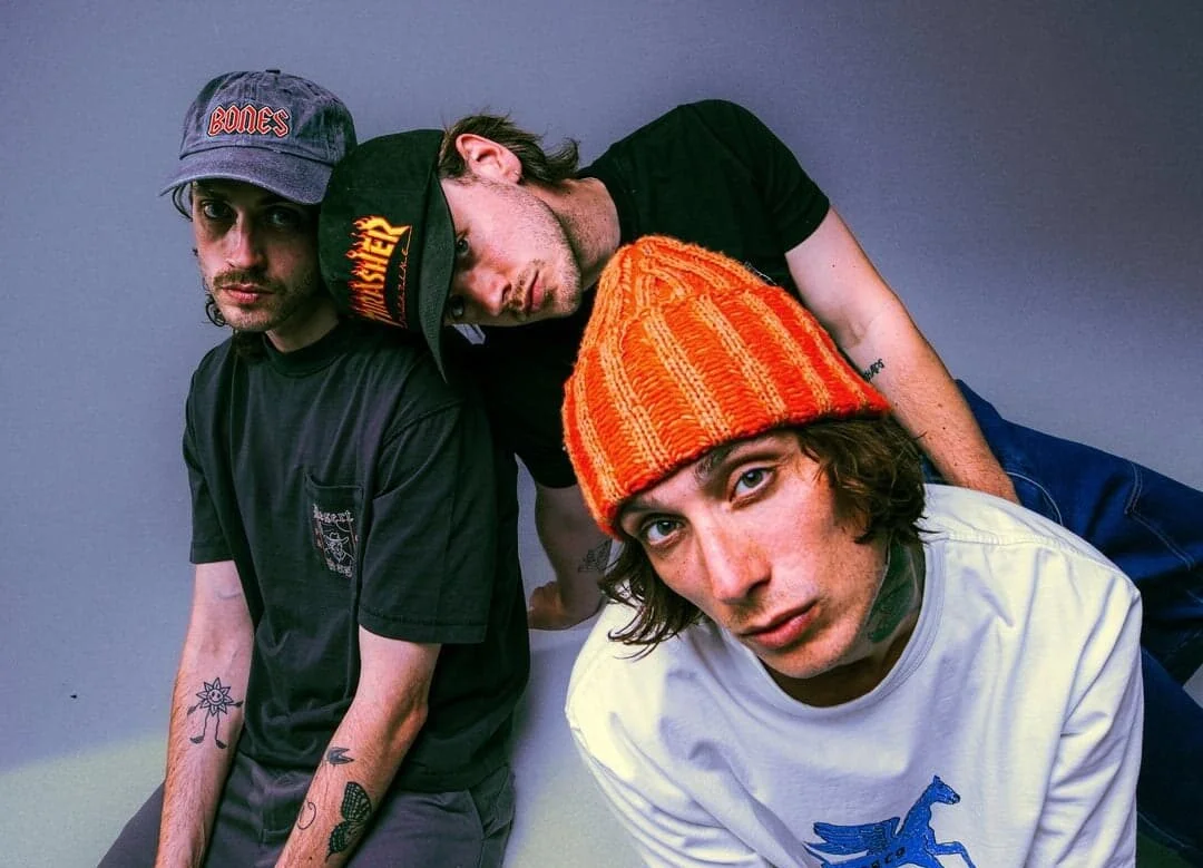 Three young men with casual clothing and hats, posing closely together against a plain background.