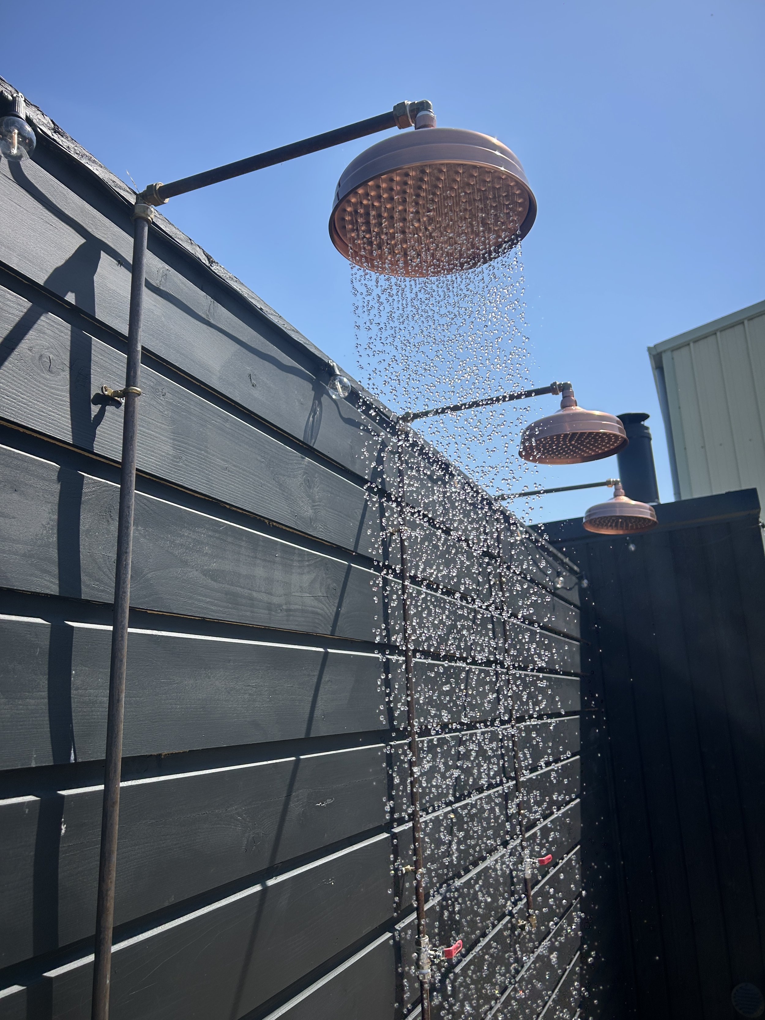 Outdoor shower head mounted on wall, water spraying downward against clear blue sky.