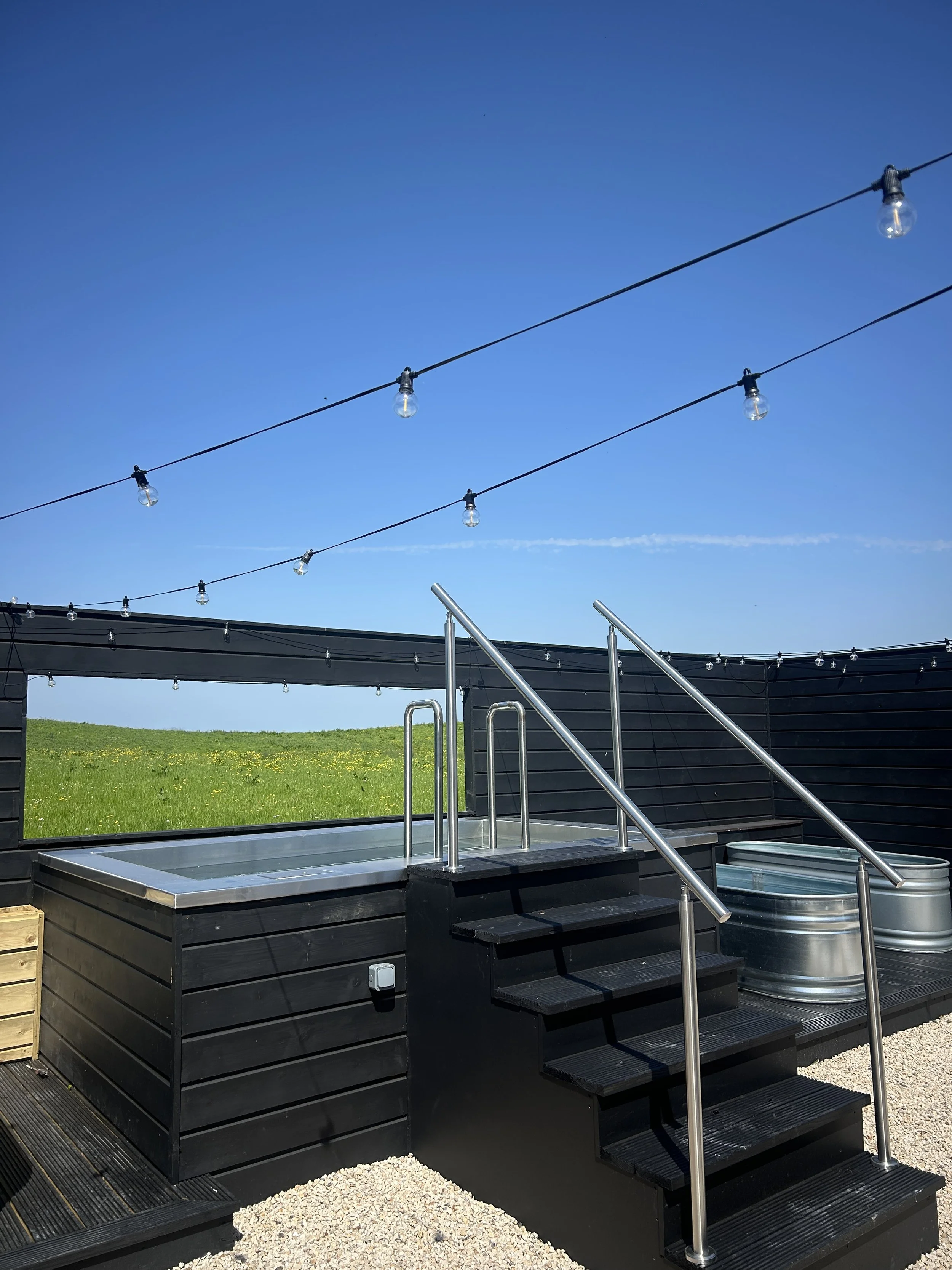 A hot tub with steps leading up to it, surrounded by a black wooden frame. Above are string lights hanging in the sky, with a clear blue sky and green grass in the background.