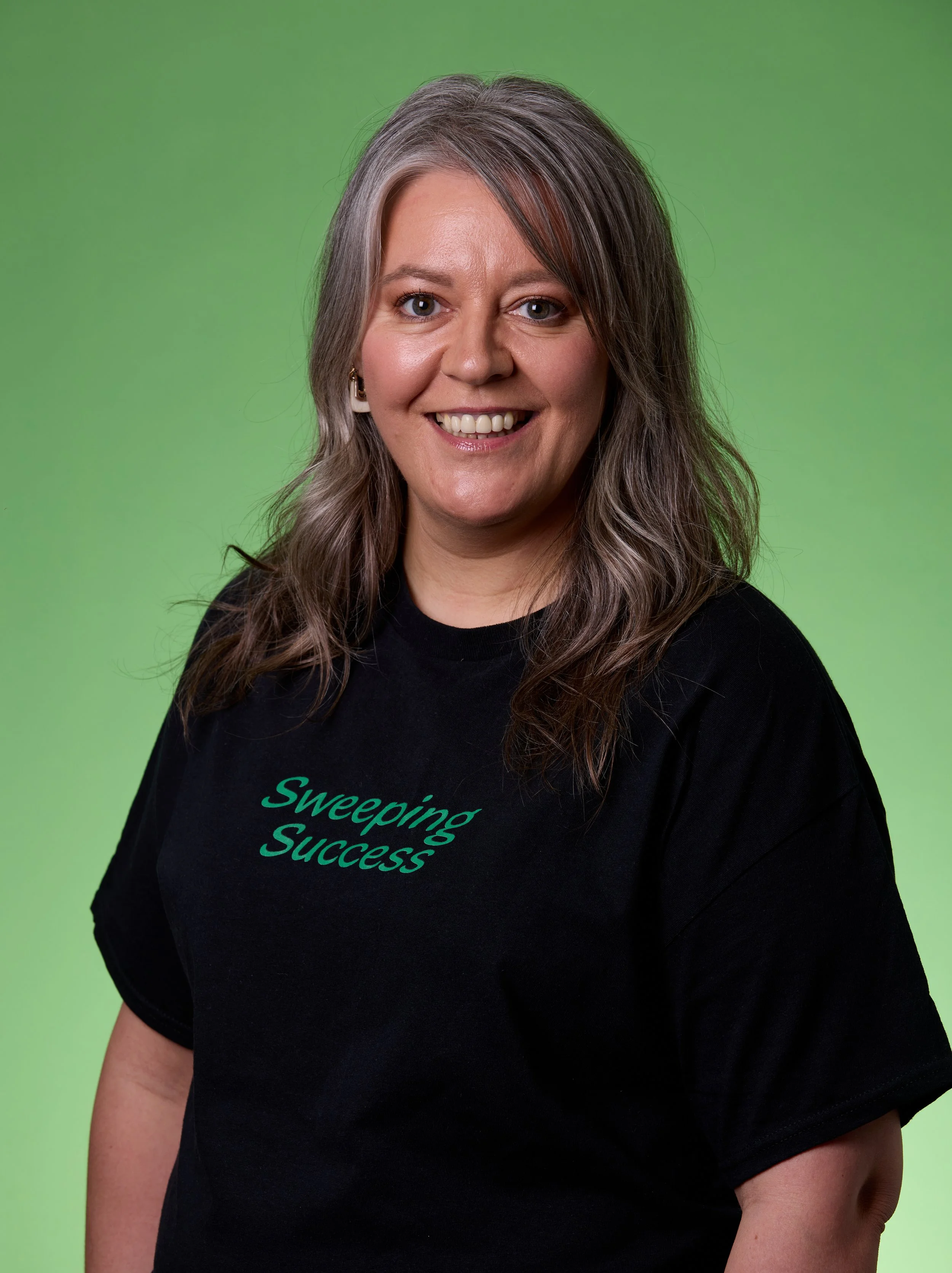 A woman with shoulder-length wavy gray hair smiling, wearing a black T-shirt with green text that says "Sweeping Success," against a green background.