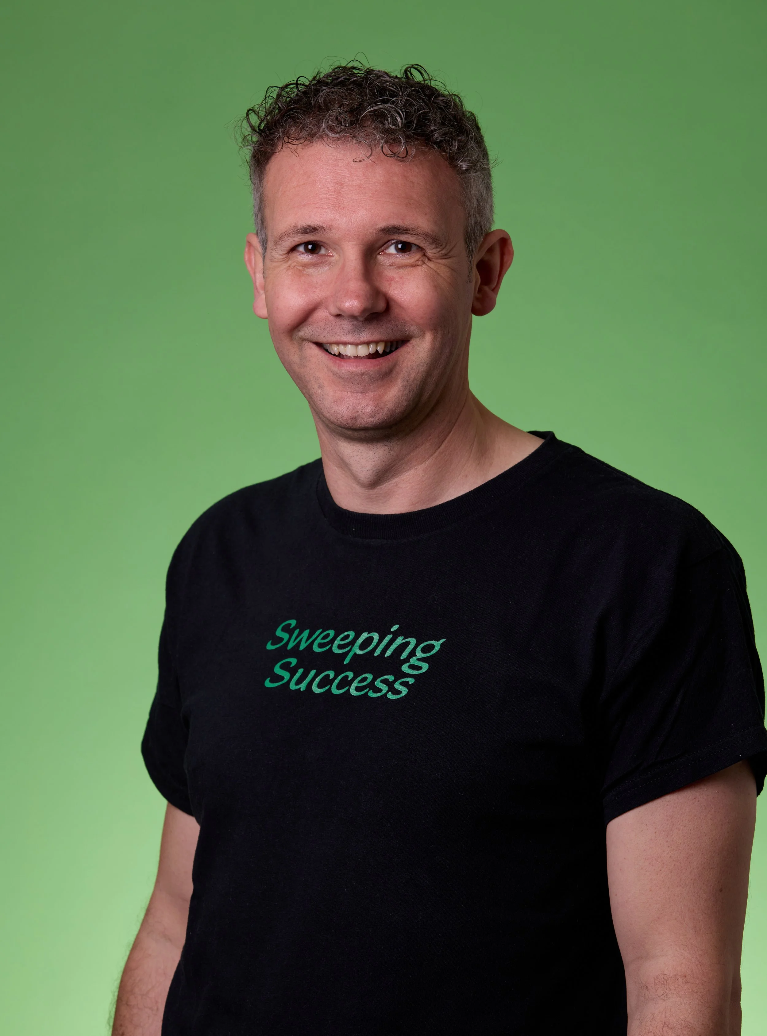 A man with curly hair and a smile, wearing a black t-shirt that says 'Sweeping Success', standing against a green background.