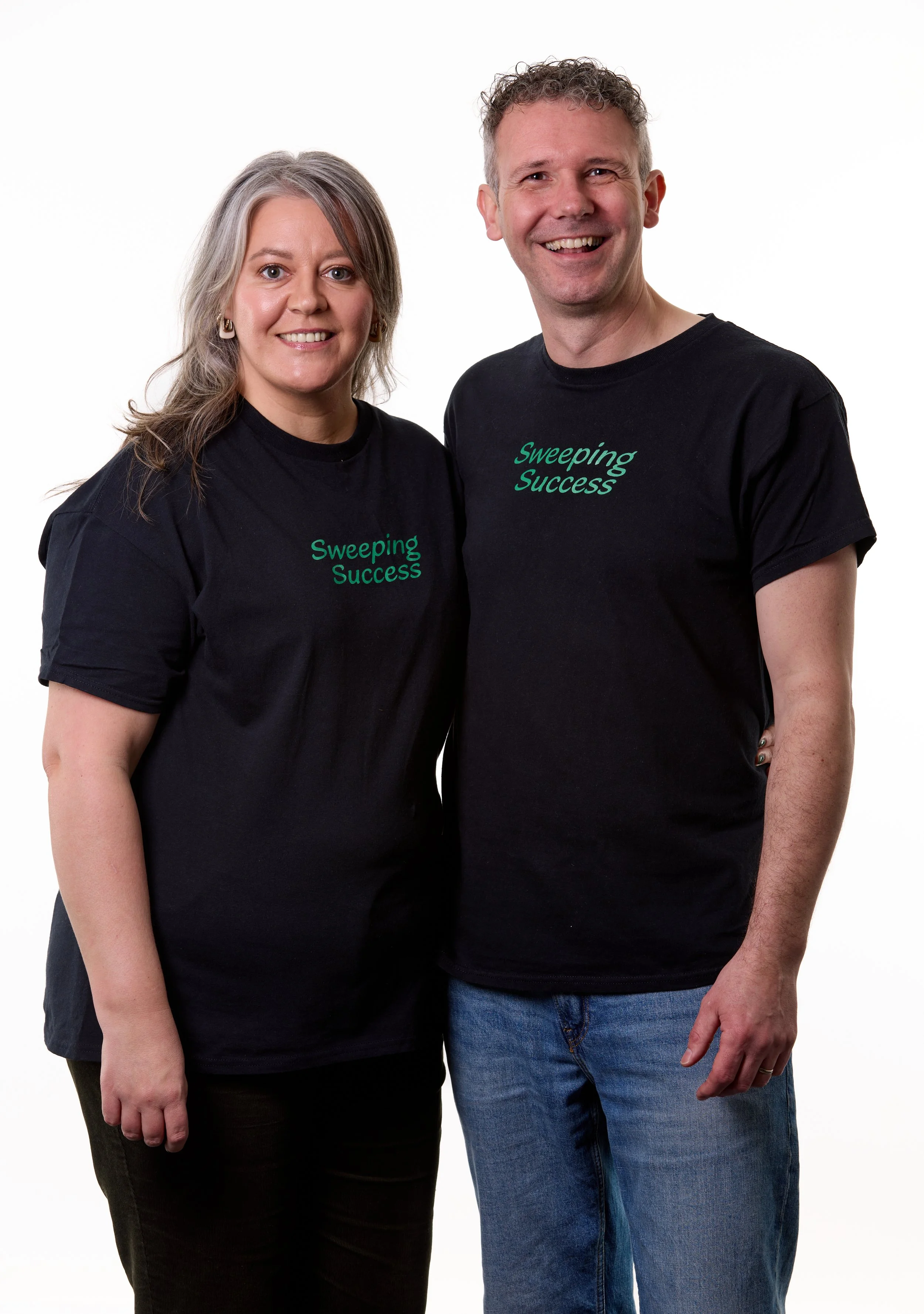 A woman and a man smiling while standing together against a white background, both wearing black T-shirts with green text that reads 'Sweeping Success'.