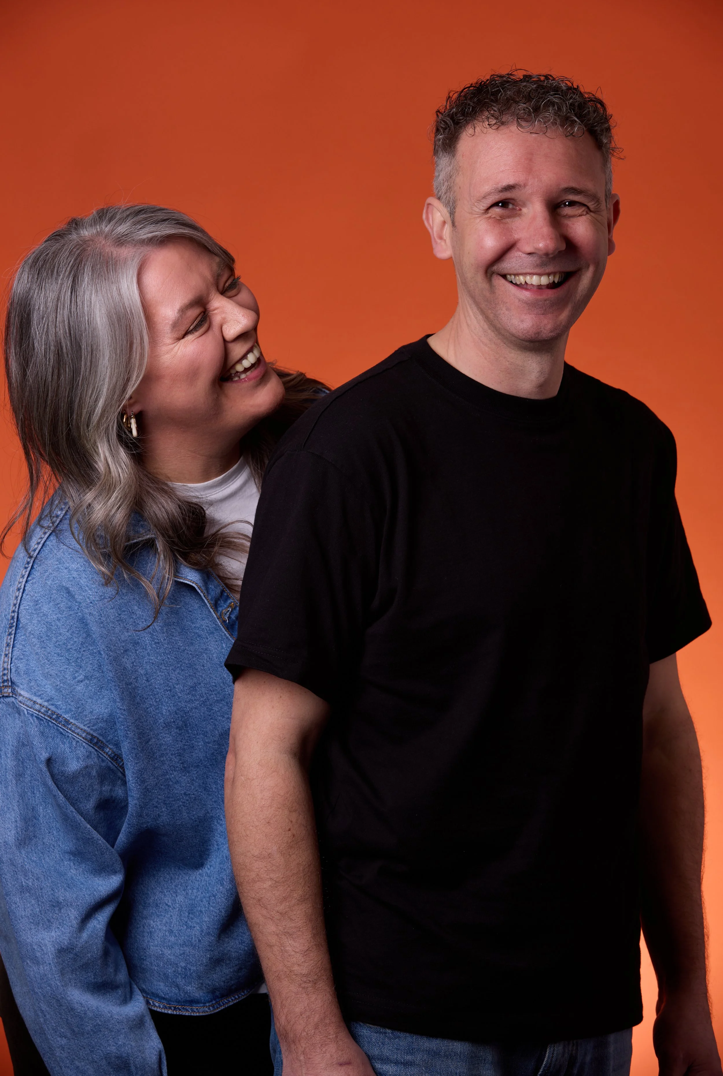 A smiling woman with gray hair leaning on a smiling man with curly hair against an orange background.