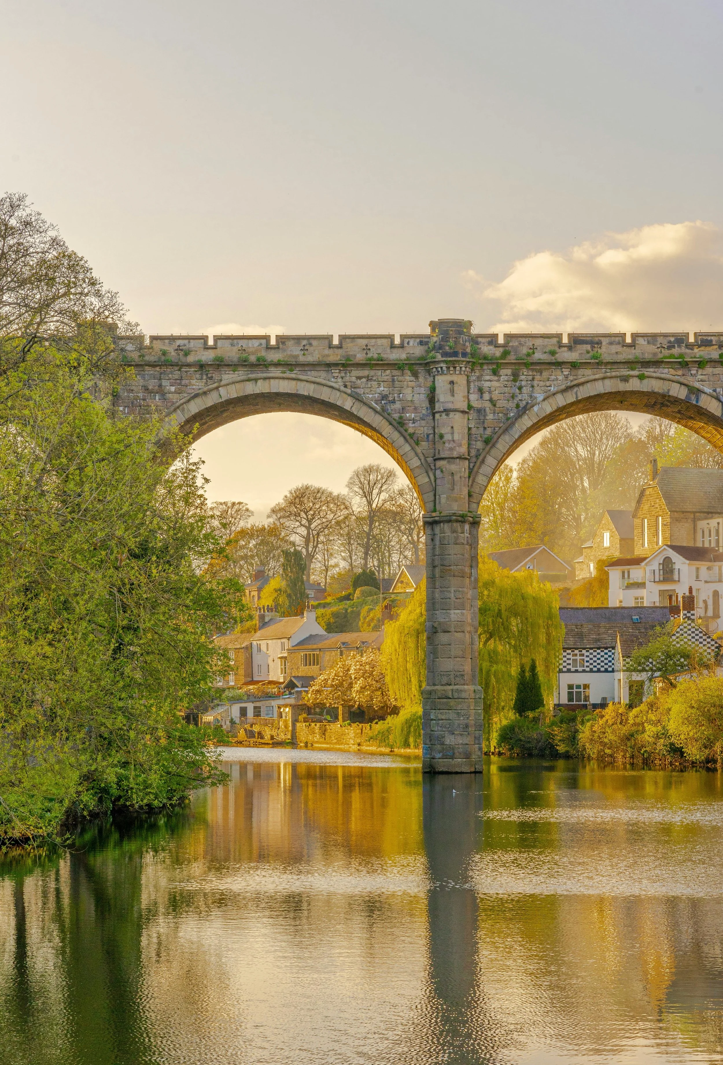 Knaresborough. A stone arch bridge over a river with houses and trees along the banks, during golden hour with warm sunlight.