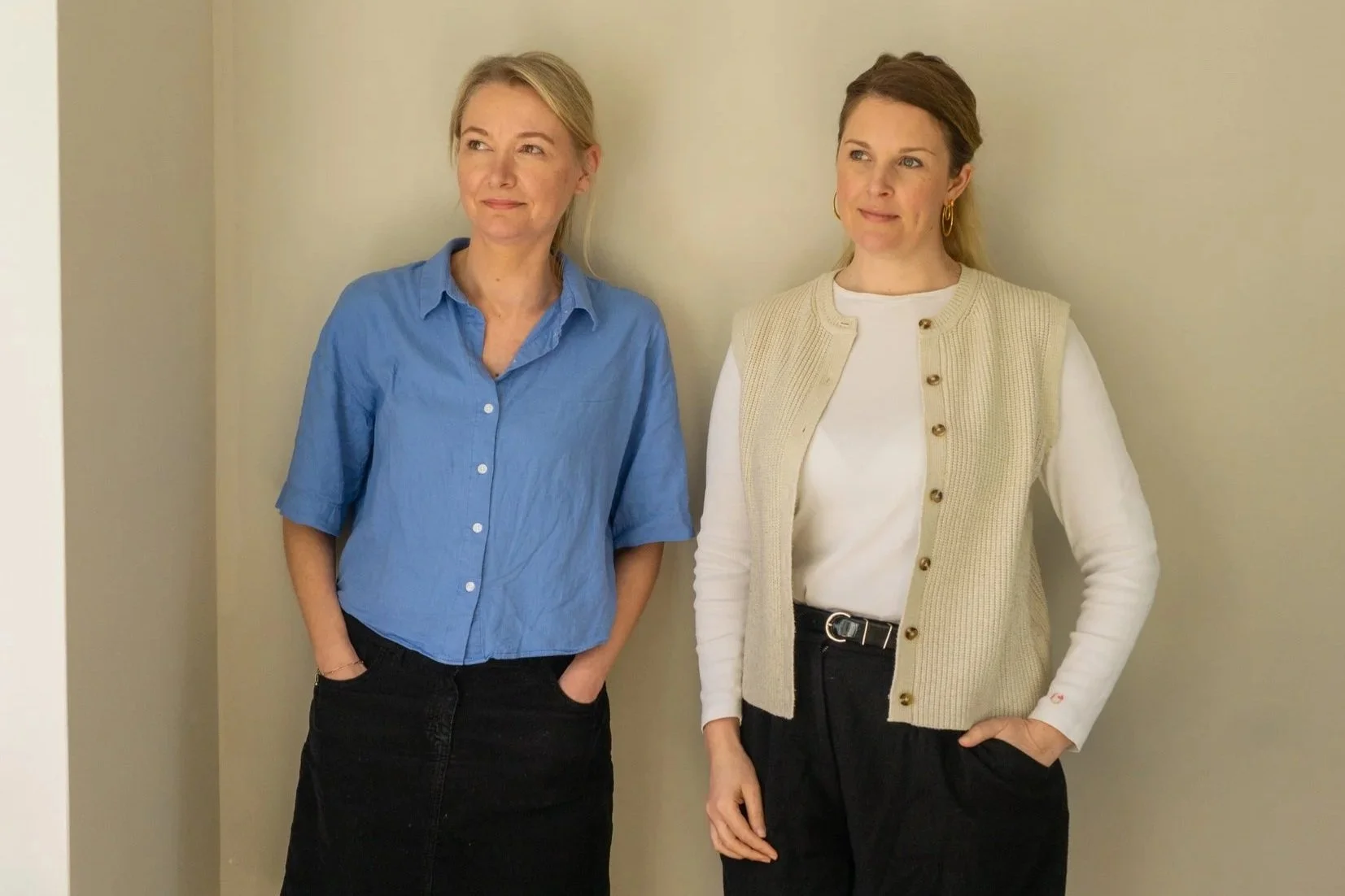 Polly Woodvine and Melanie Hill standing against a beige wall, one in a blue shirt and black skirt, the other in a white shirt with a beige vest, both with their hands in their pockets, looking thoughtfully to the side.