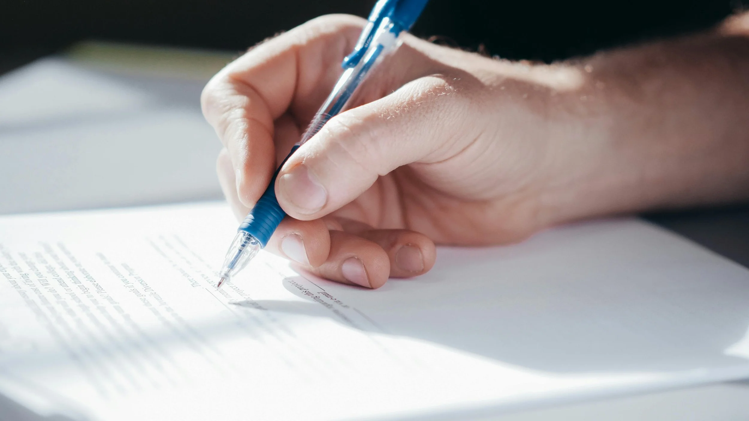 Close-up of a hand writing with a blue pen on white paper in natural light.