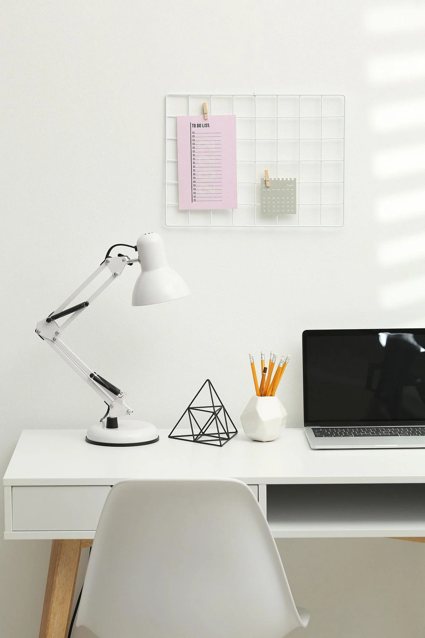 Minimalist white desk with a white adjustable lamp, geometric wire sculpture, white vase with pencils, and a black laptop against a white wall with a grid note board.