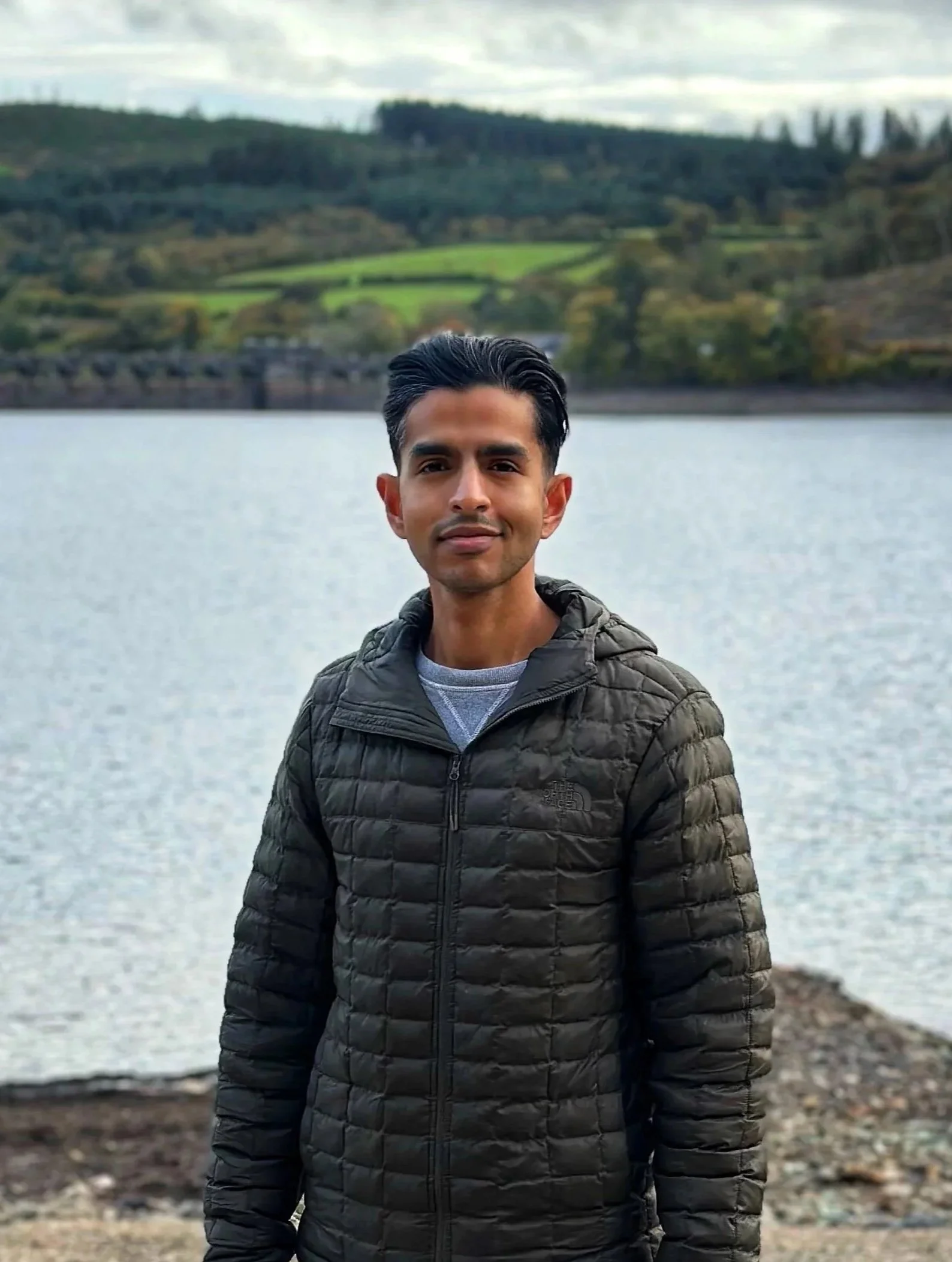 A young man stands by a lake with hills and trees in the background, wearing a green puffer jacket. Therapist in Liverpool.