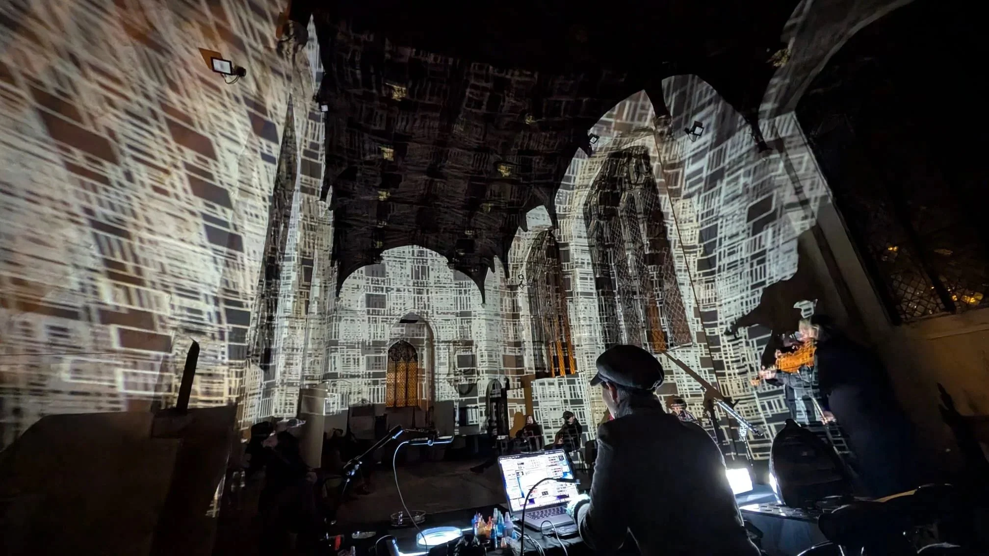 An indoor event with a projected cityscape on the walls and ceiling, people facing a stage or DJ booth with equipment and screens.