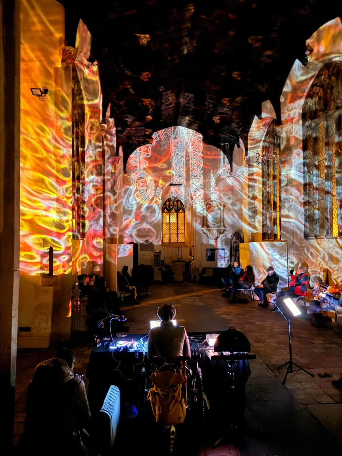 People sitting on chairs in a historic church with colorful light projections on the walls and ceiling during a nighttime event, with a DJ at the front.