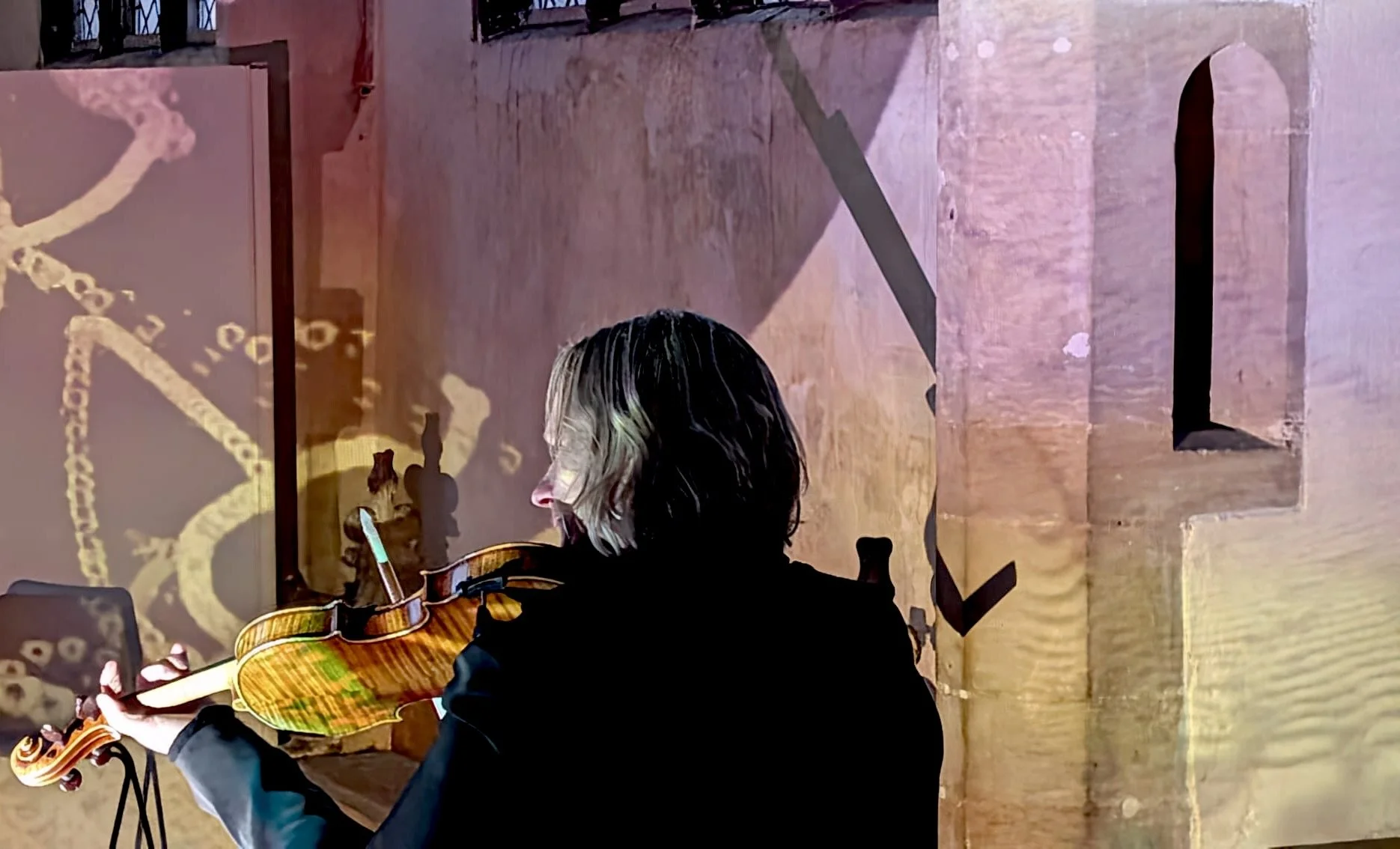 Person with shoulder-length hair playing an acoustic guitar during a performance or practice session, with colorful lighting and shadows on the wall.