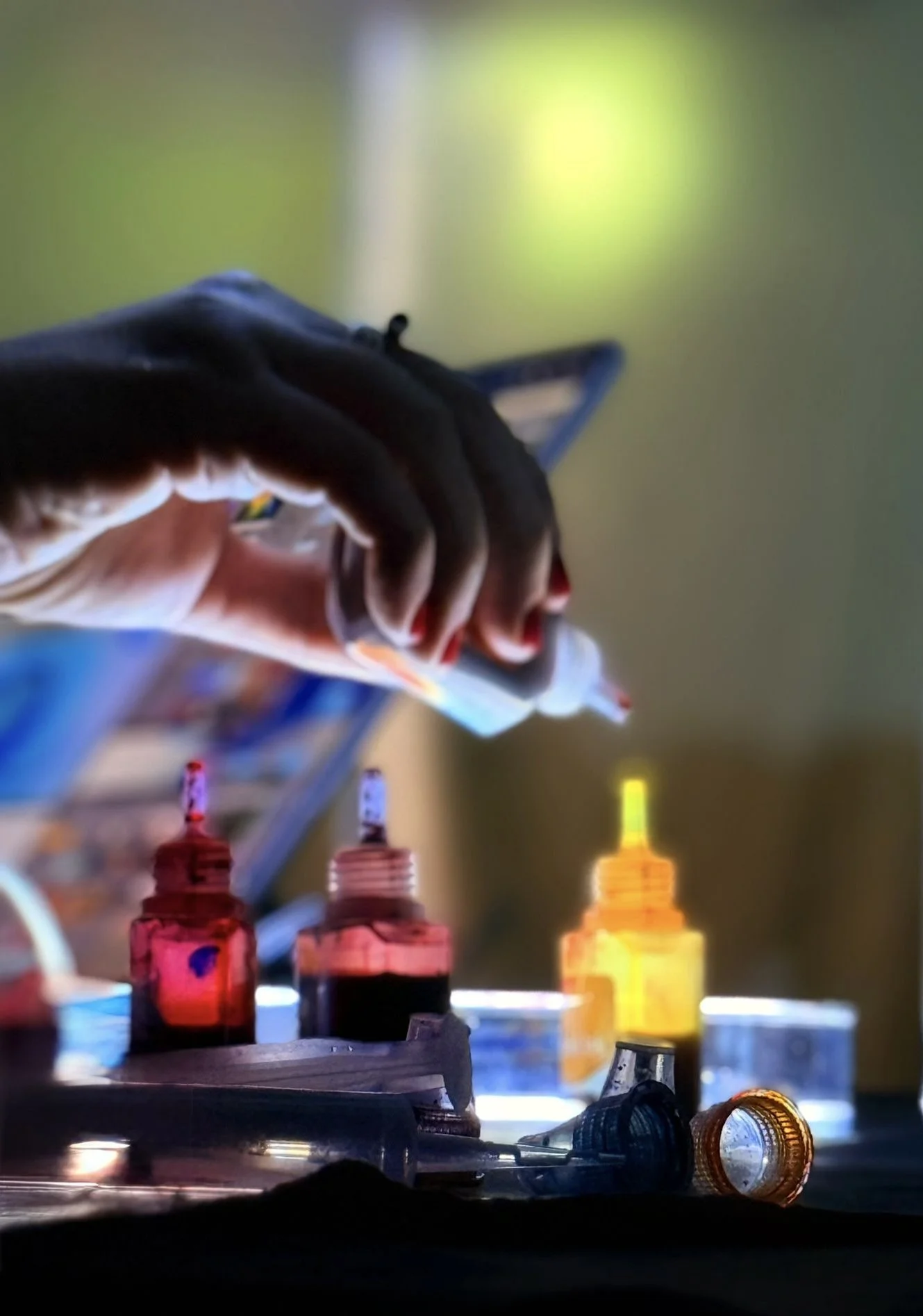Close-up of paint bottles and a hand holding a paintbrush in front of a blurred background, possibly in an art studio.