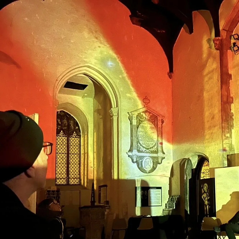 An interior view of a church or cathedral with colorful light projections on the walls, creating a vibrant atmosphere. A person with glasses is partially visible on the left.