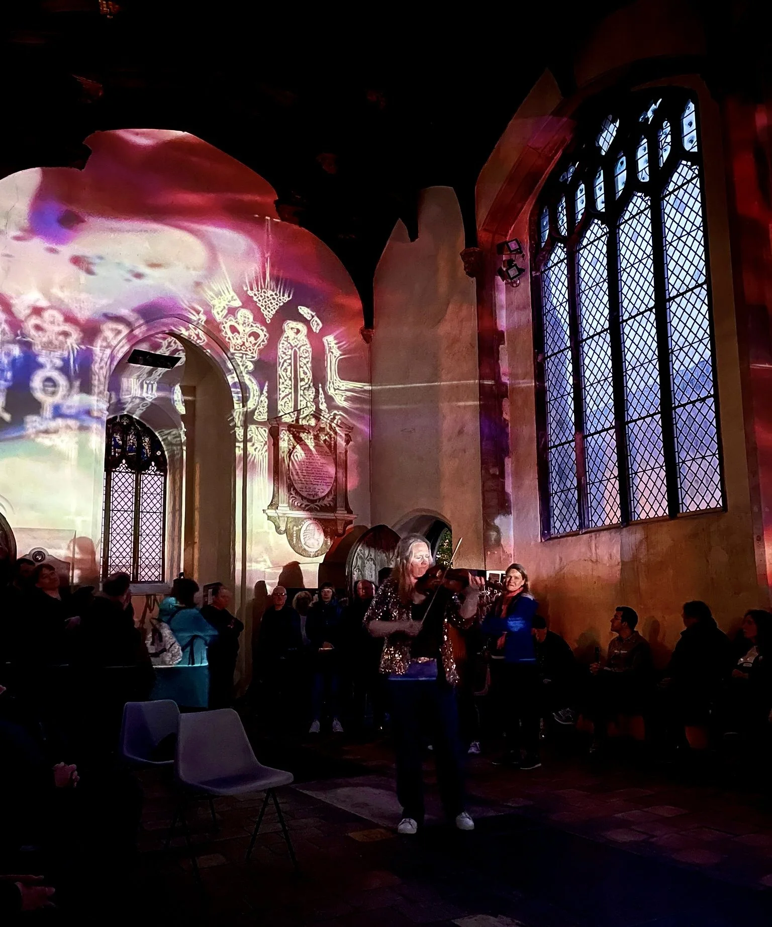 A group of people inside a dimly lit church or chapel with large stained glass windows, where a woman is playing the violin while others sit and stand around, and colorful light projections are displayed on the walls.