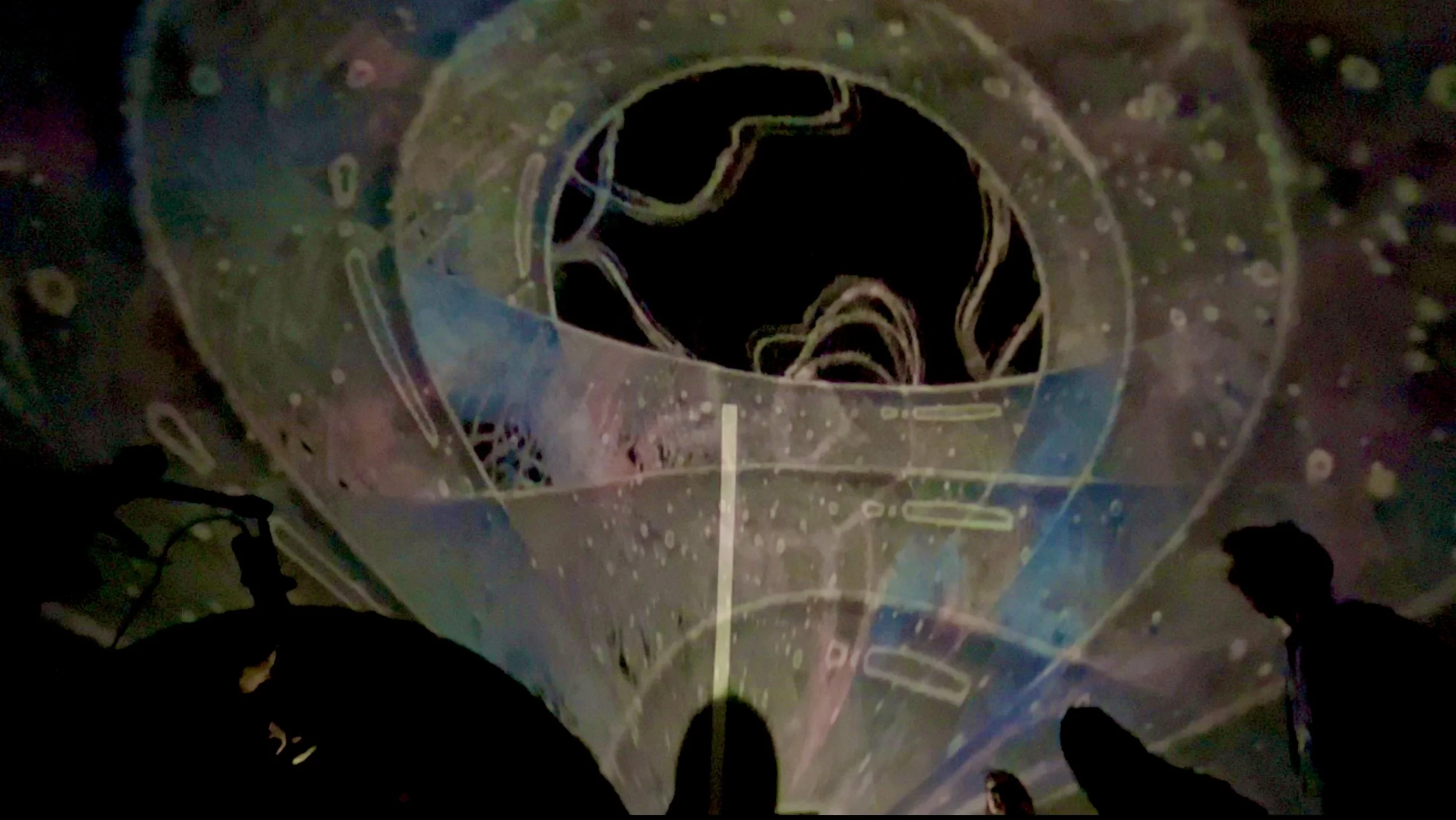 A large donut-shaped structure with a black interior and neon-colored outline, surrounded by smaller objects and people in a dark setting.