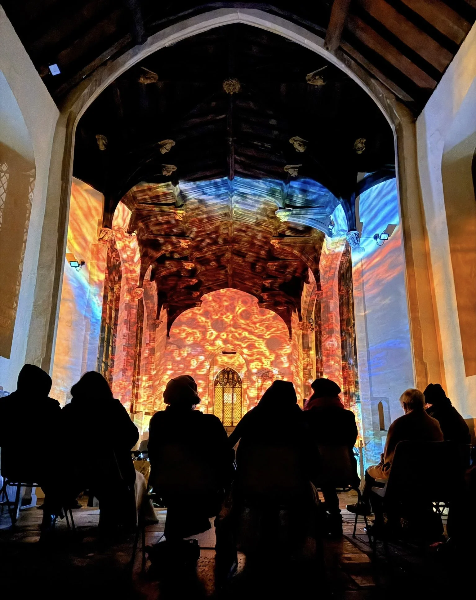 People are seated inside a church looking at a colorful, illuminated digital projection on the altar and walls, depicting a vibrant fiery scene.