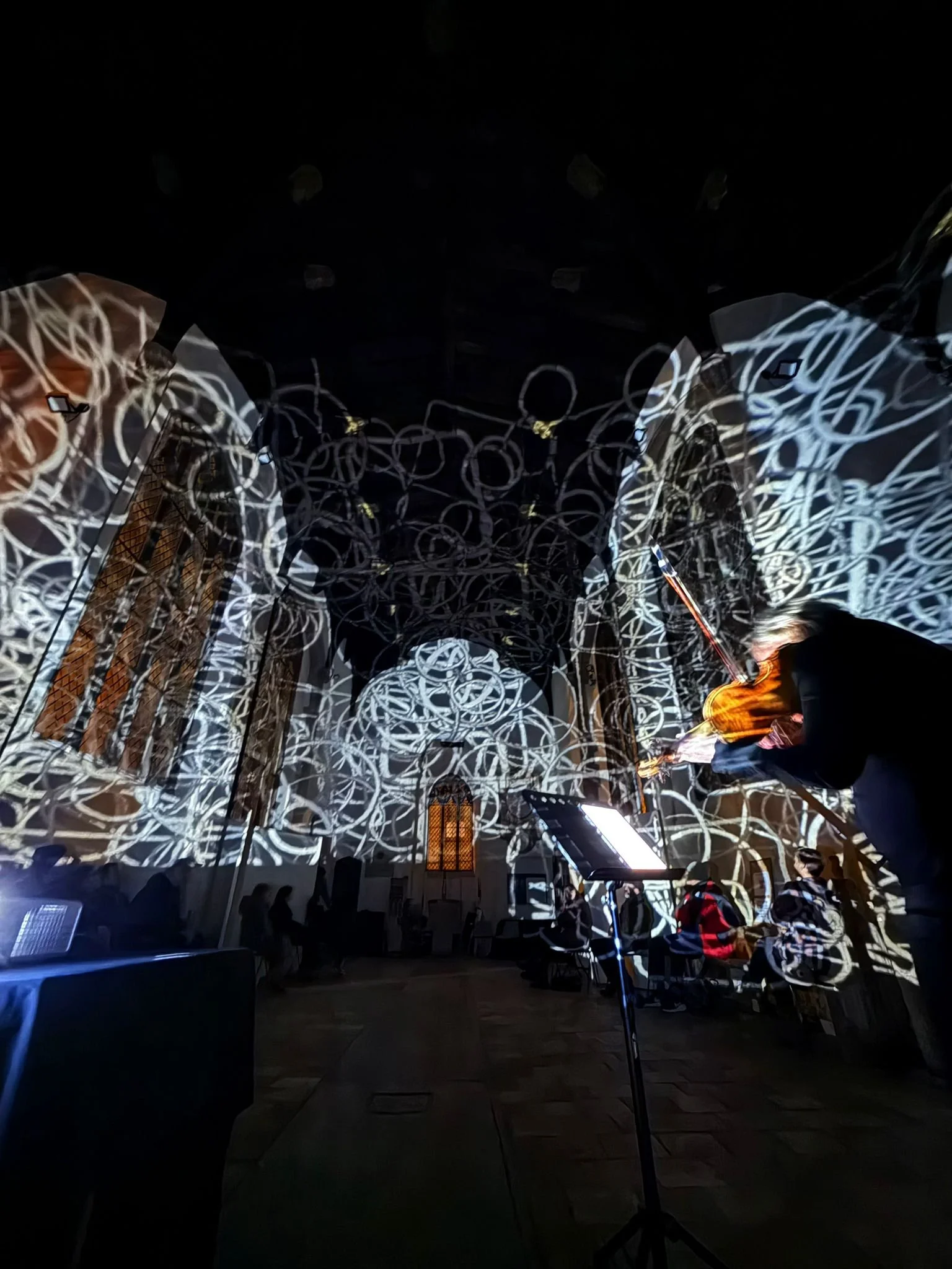 People sitting and standing in a dark room with a ceiling and walls illuminated by projections of white, circular, interconnected patterns. Some individuals focus on electronic devices, with one person using a laptop, and there is a music stand with 