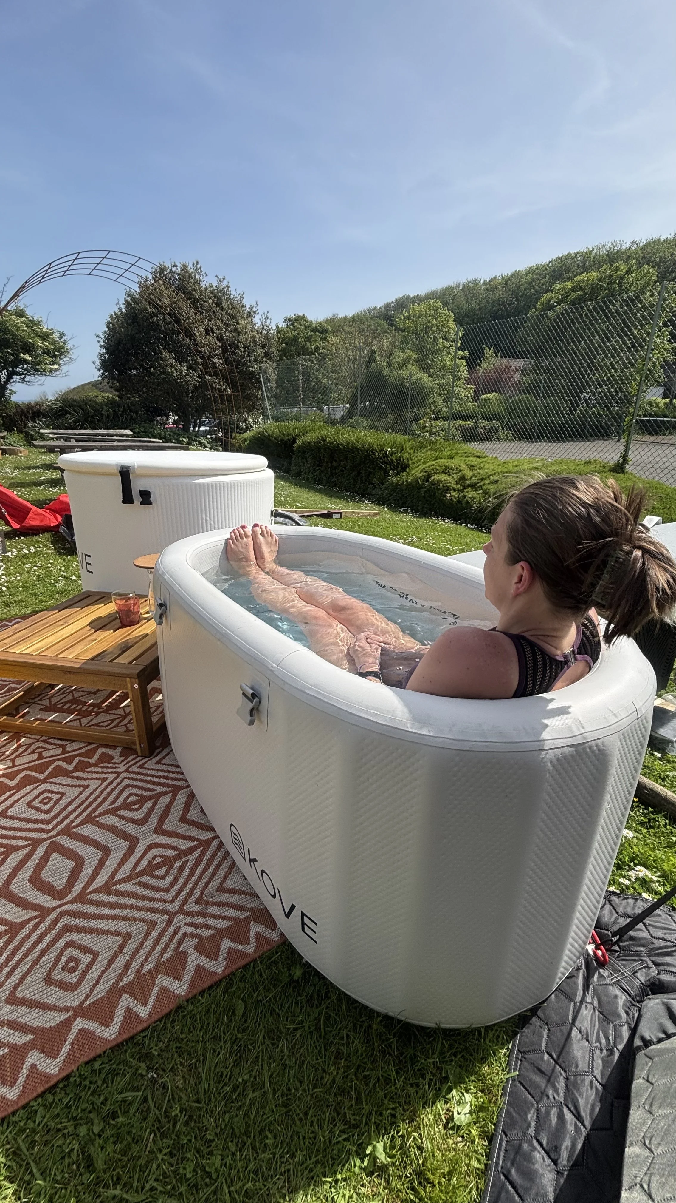 A woman relaxing in an inflatable hot tub outdoors on a grassy area with trees and a chain-link fence in the background. A small wooden table with a drink on it is beside the hot tub, and a patterned rug is underneath.