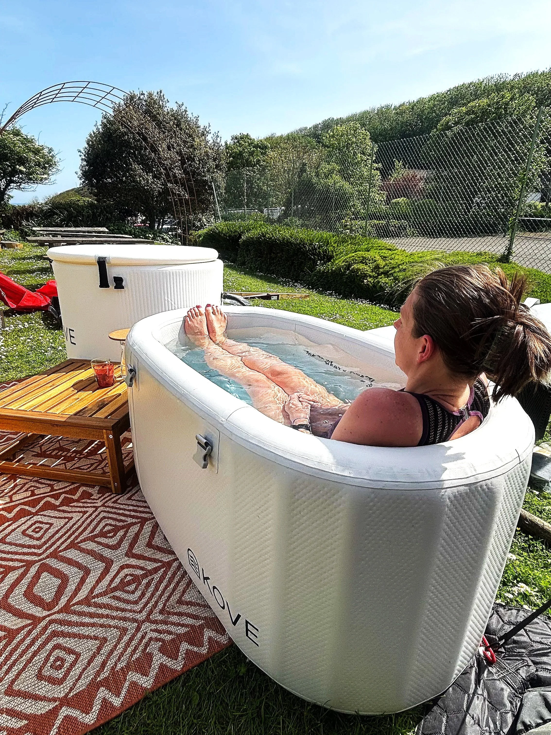 Person relaxing in a small white inflatable pool outdoors on a sunny day, with trees in the background. There is a patterned outdoor rug underneath the pool and a small wooden table with a glass of drink nearby.