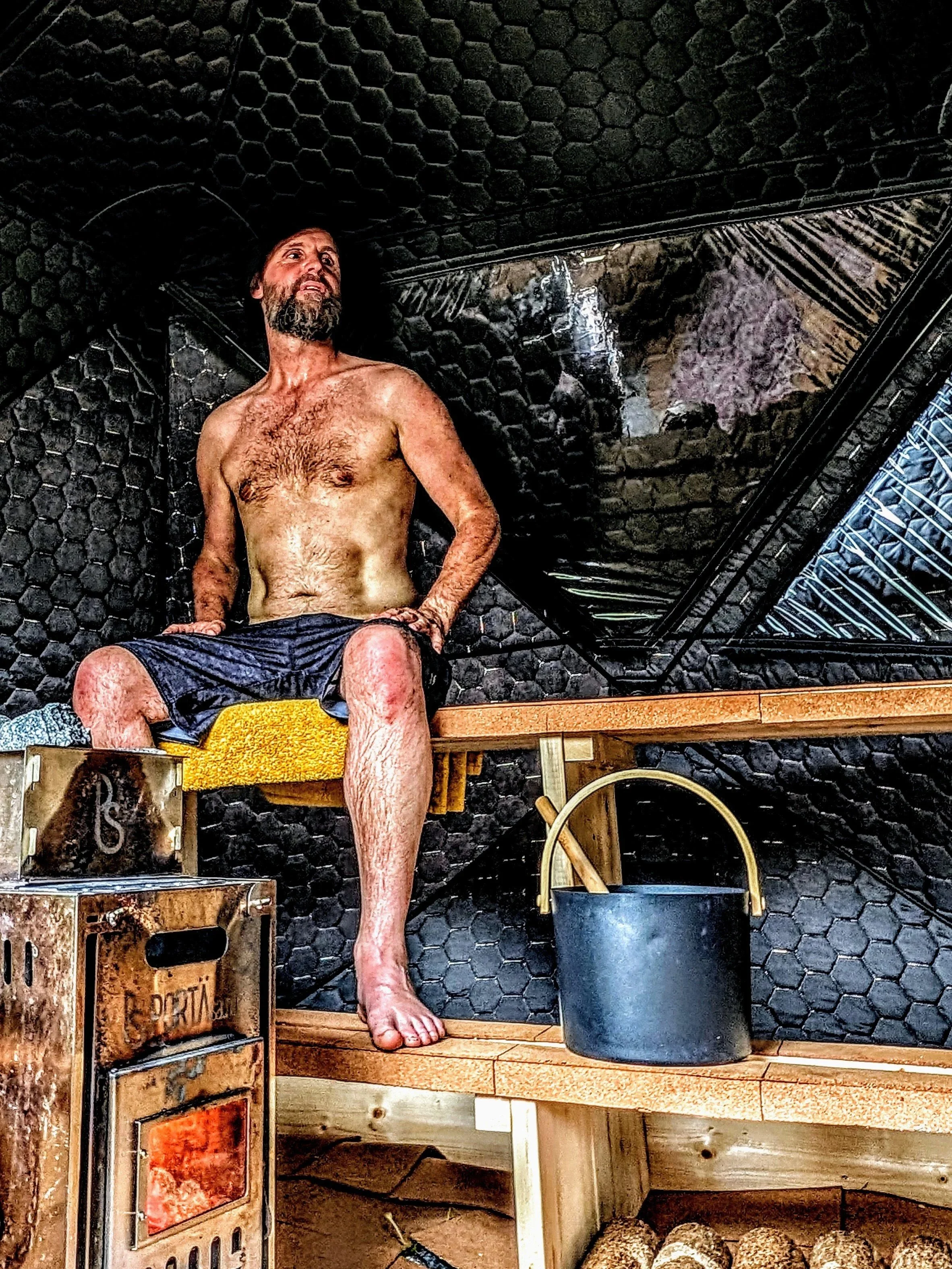 A shirtless man with a beard sitting on a wooden bench inside a sauna, with black walls and a small stove in front of him. There are wooden benches, buckets, and logs nearby.
