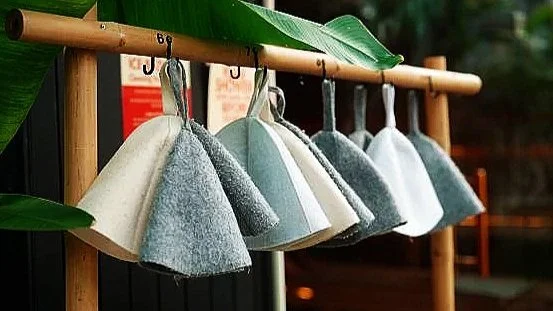 Felt sauna hats hanging on a wooden rack outdoors with green leaves nearby.