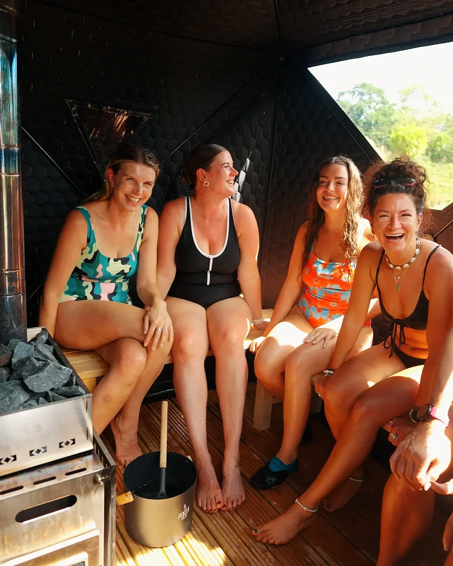 Four women sitting inside a sauna or steam room, smiling and laughing together, dressed in swimsuits.