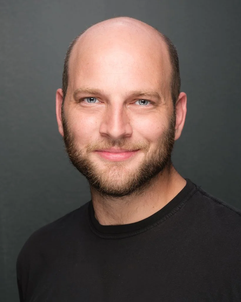A close-up portrait of a smiling man with a beard and blue eyes, wearing a black shirt, against a gray background.