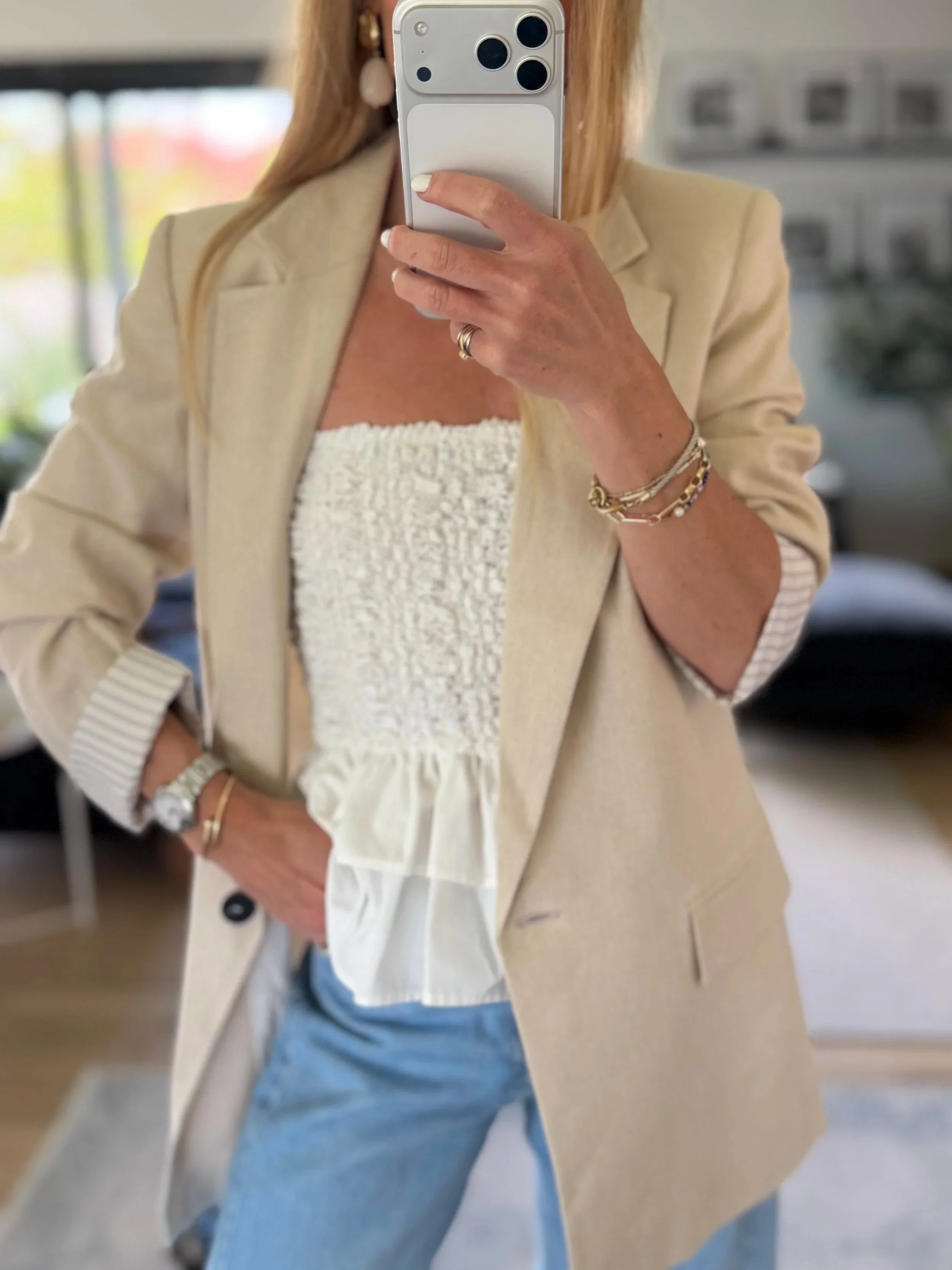 A woman taking a mirror selfie showing her beige blazer, white top, blue jeans, and jewelry including bracelets and rings.