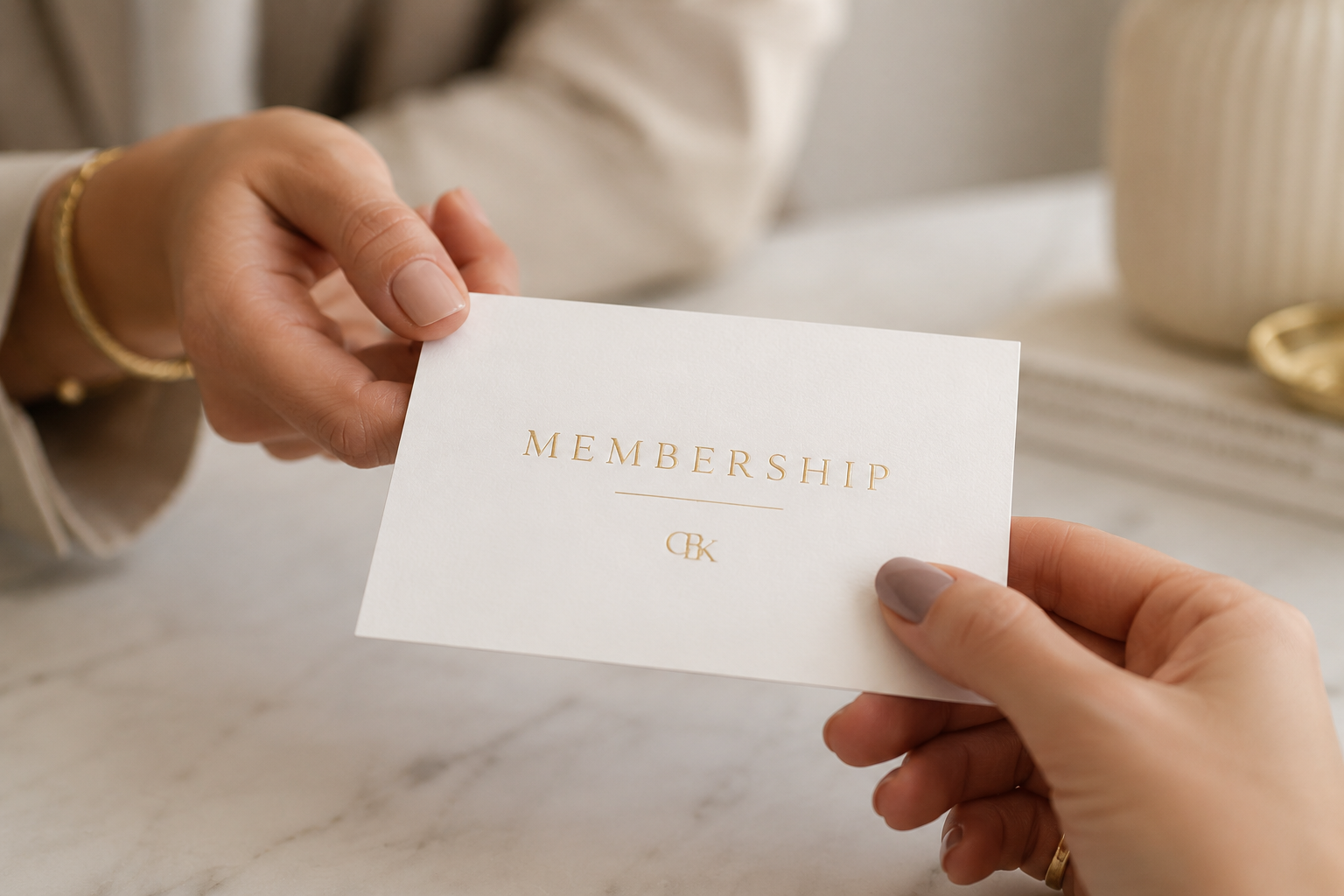 Two hands holding a white card that says 'Membership' with gold lettering, with a blurred background including a gold bracelet and a beige container.