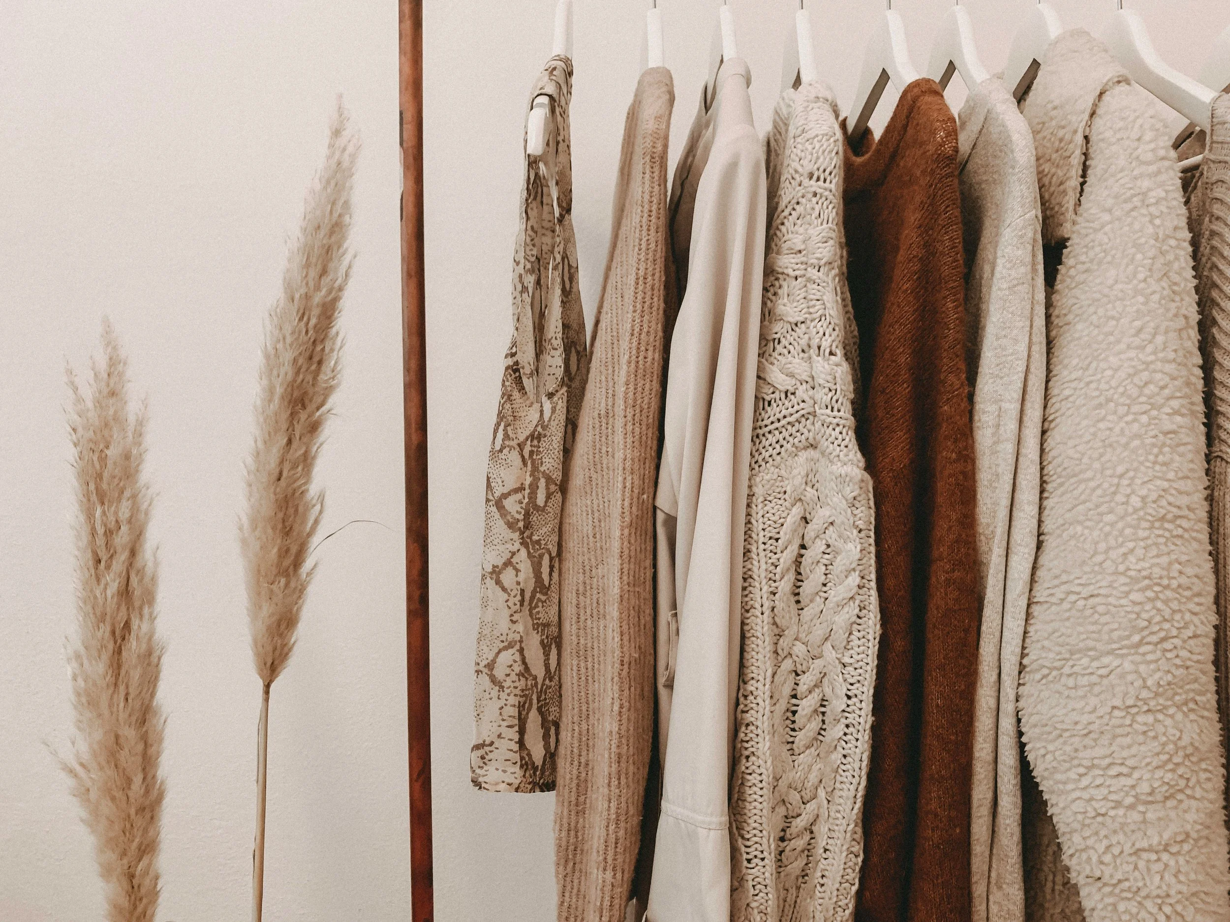 A clothing rack with various beige, white, and brown sweaters and shirts, and beige pampas grass in the background.