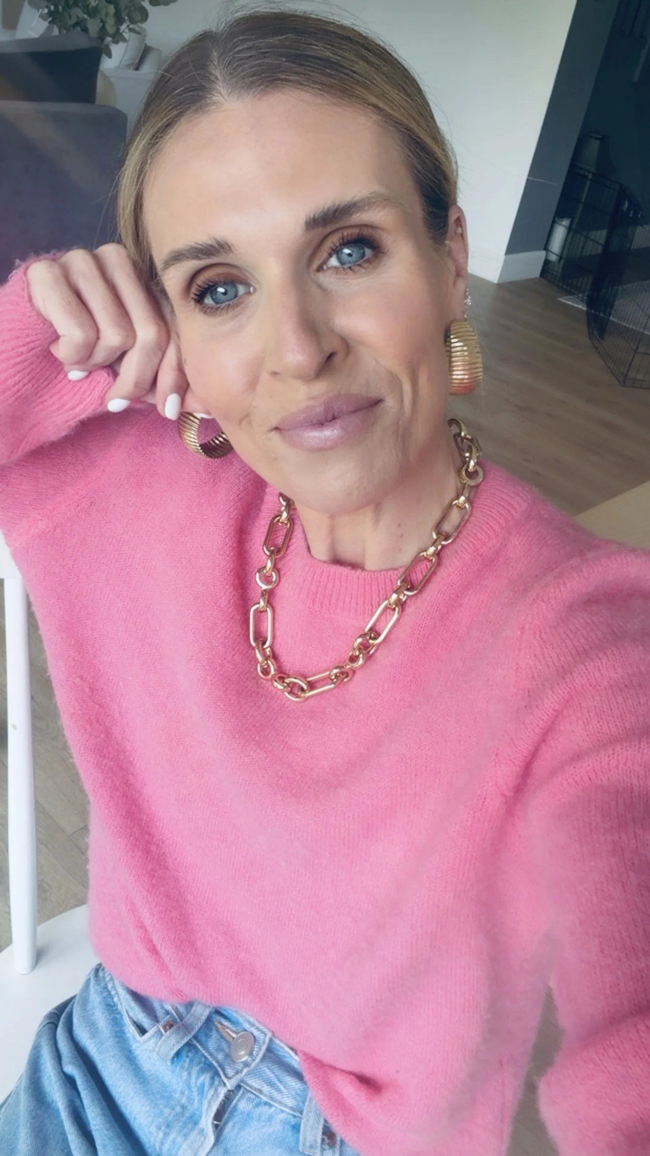 Close-up of a woman with blue eyes, wearing a pink sweater, gold chain necklace, and large gold earrings, sitting indoors with a relaxed pose and hand resting on her head.