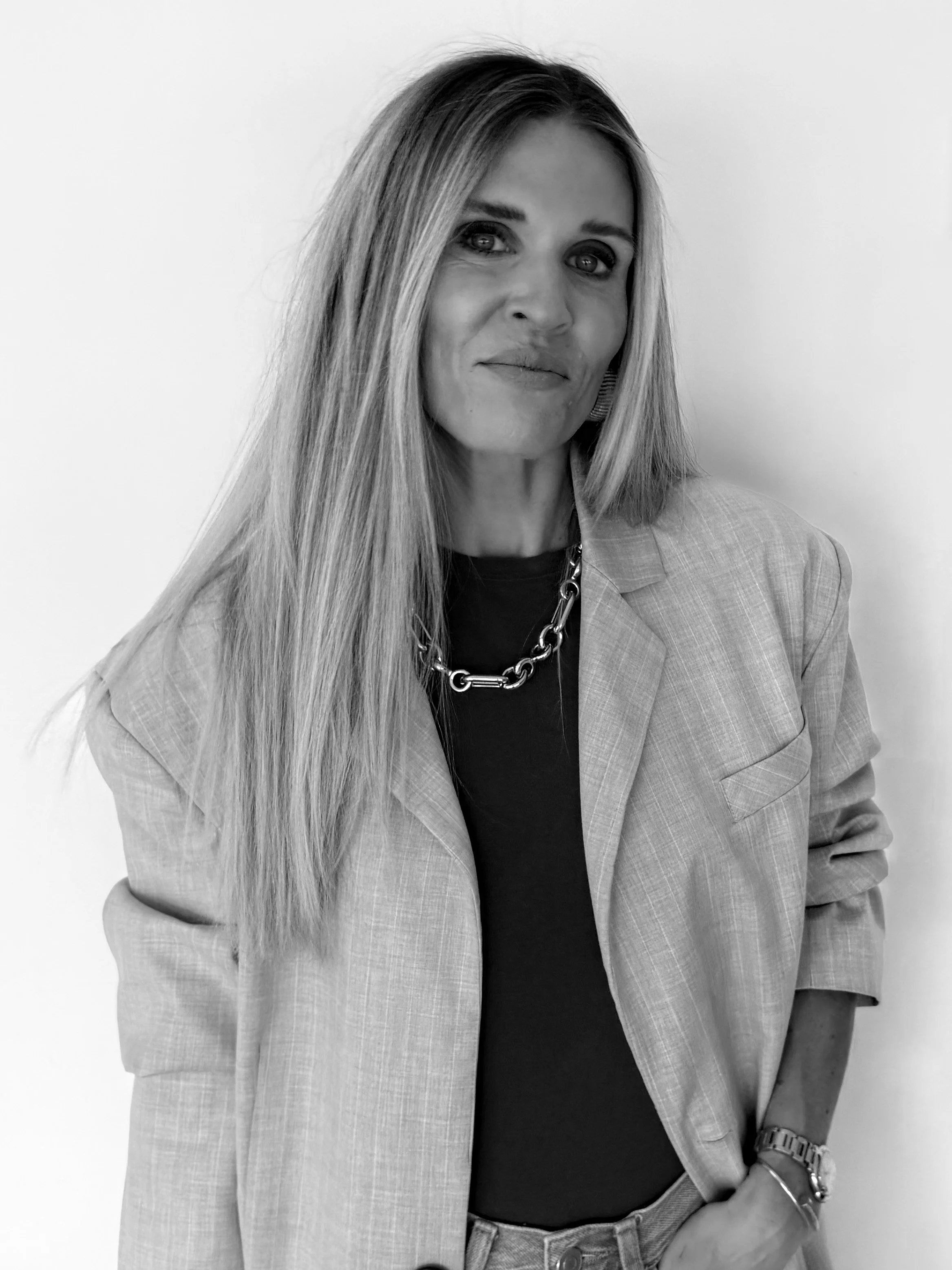 Black and white photo of a woman with long hair wearing a blazer, t-shirt, chain necklace, wristwatch, and bracelet, standing against a plain background.