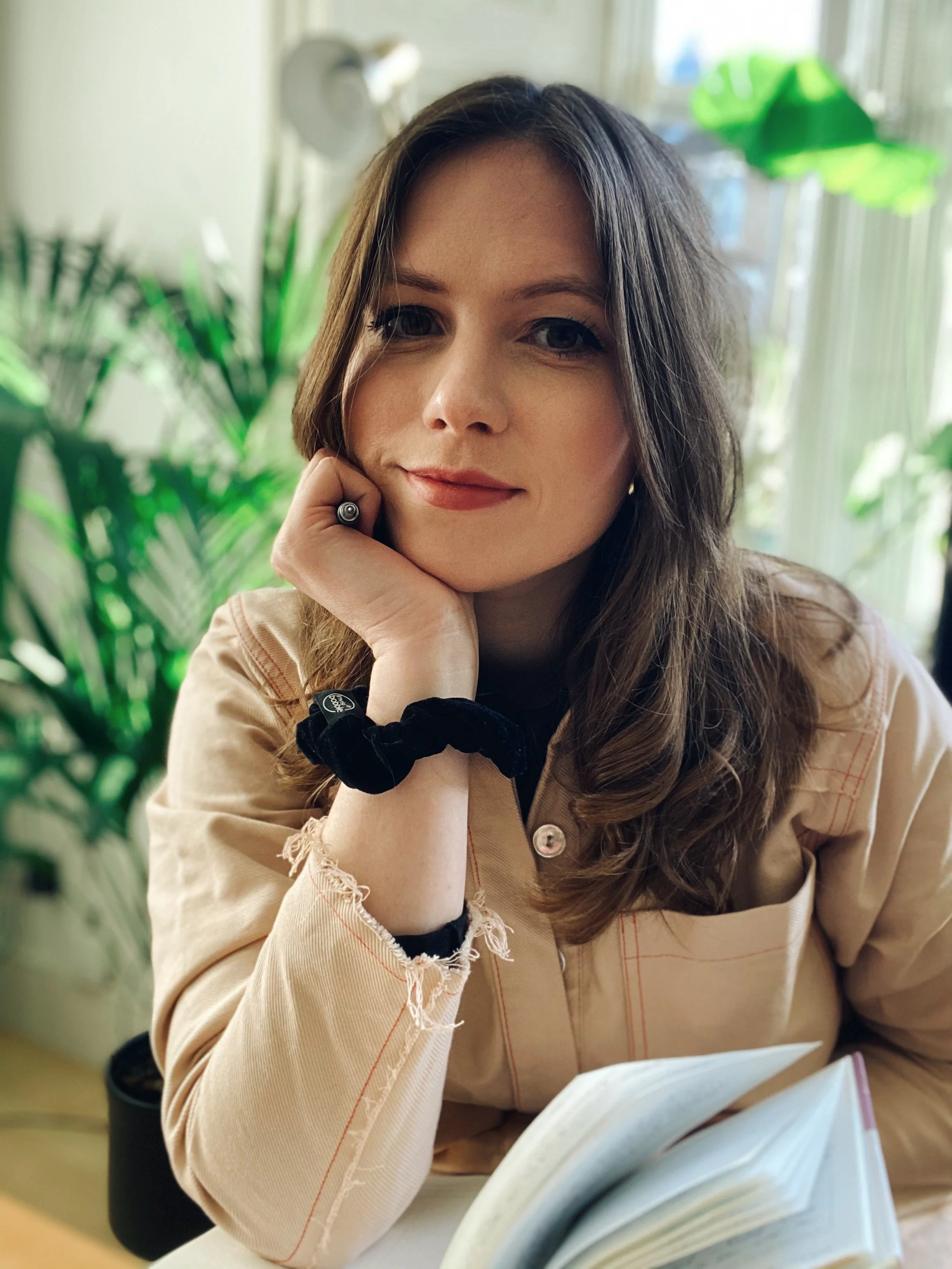 A young woman with long brown hair and fair skin, sitting indoors with natural light, resting her chin on her hand. She has a slight smile, wears a beige jacket, a black scrunchie on her wrist, and a ring on her finger. An open book is in front of her, and green plants are visible in the background.