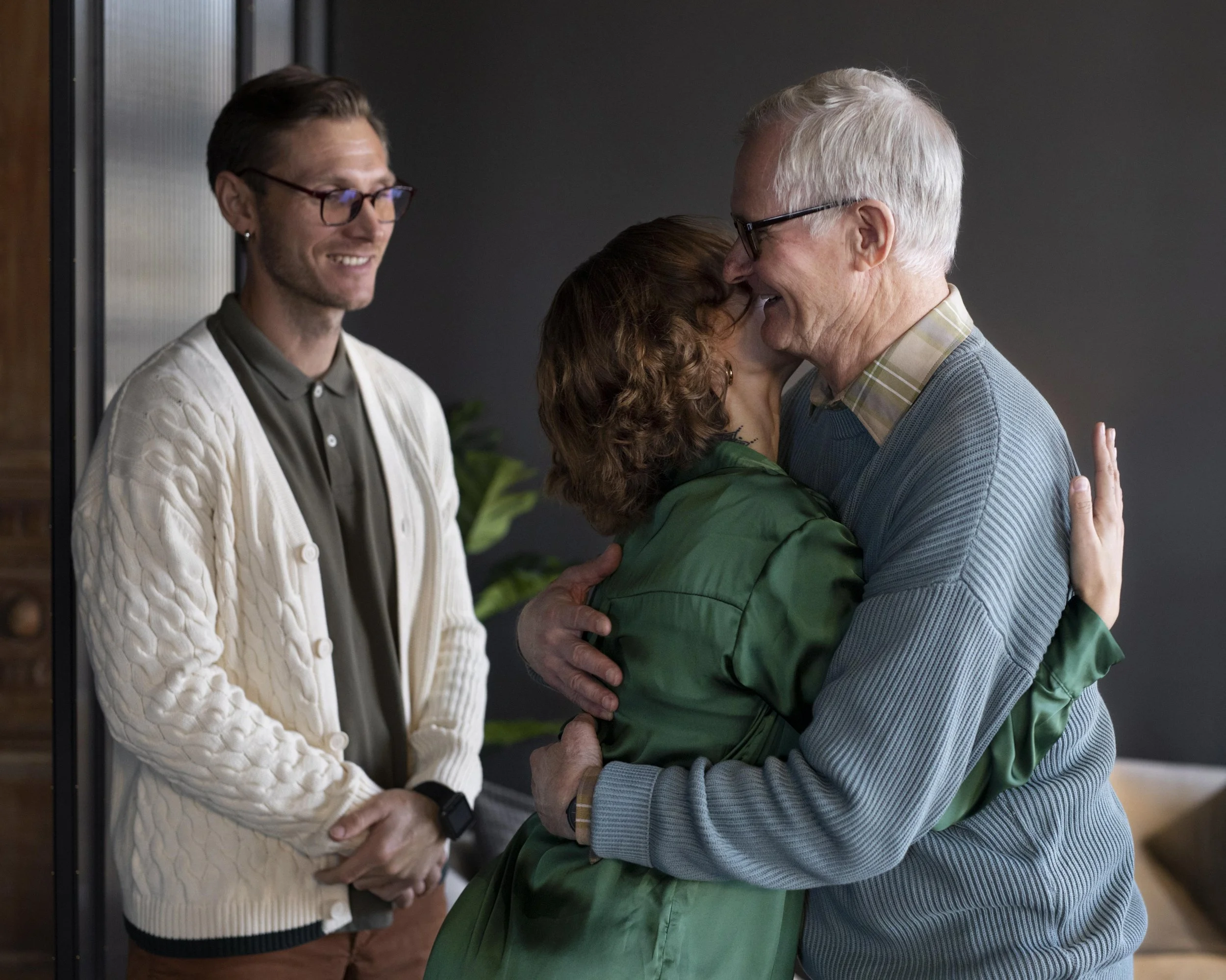 An elderly man and woman hugging, with a younger man standing nearby, smiling and observing.