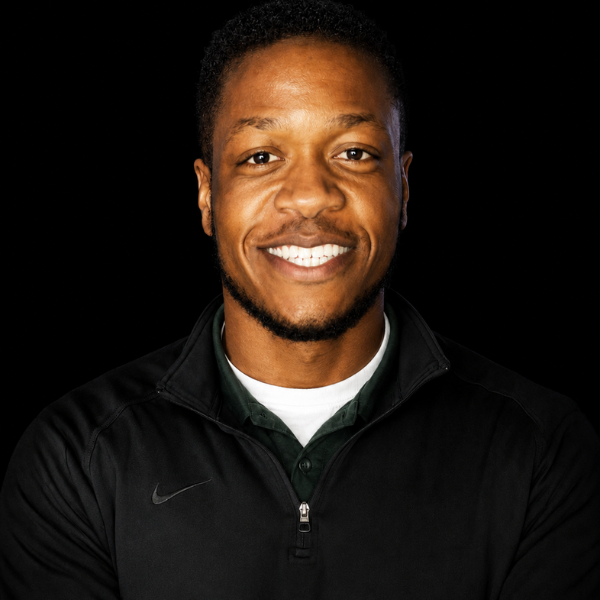 Portrait of a smiling man with short curly hair, wearing a black Nike zip-up jacket over a collared shirt, against a black background.