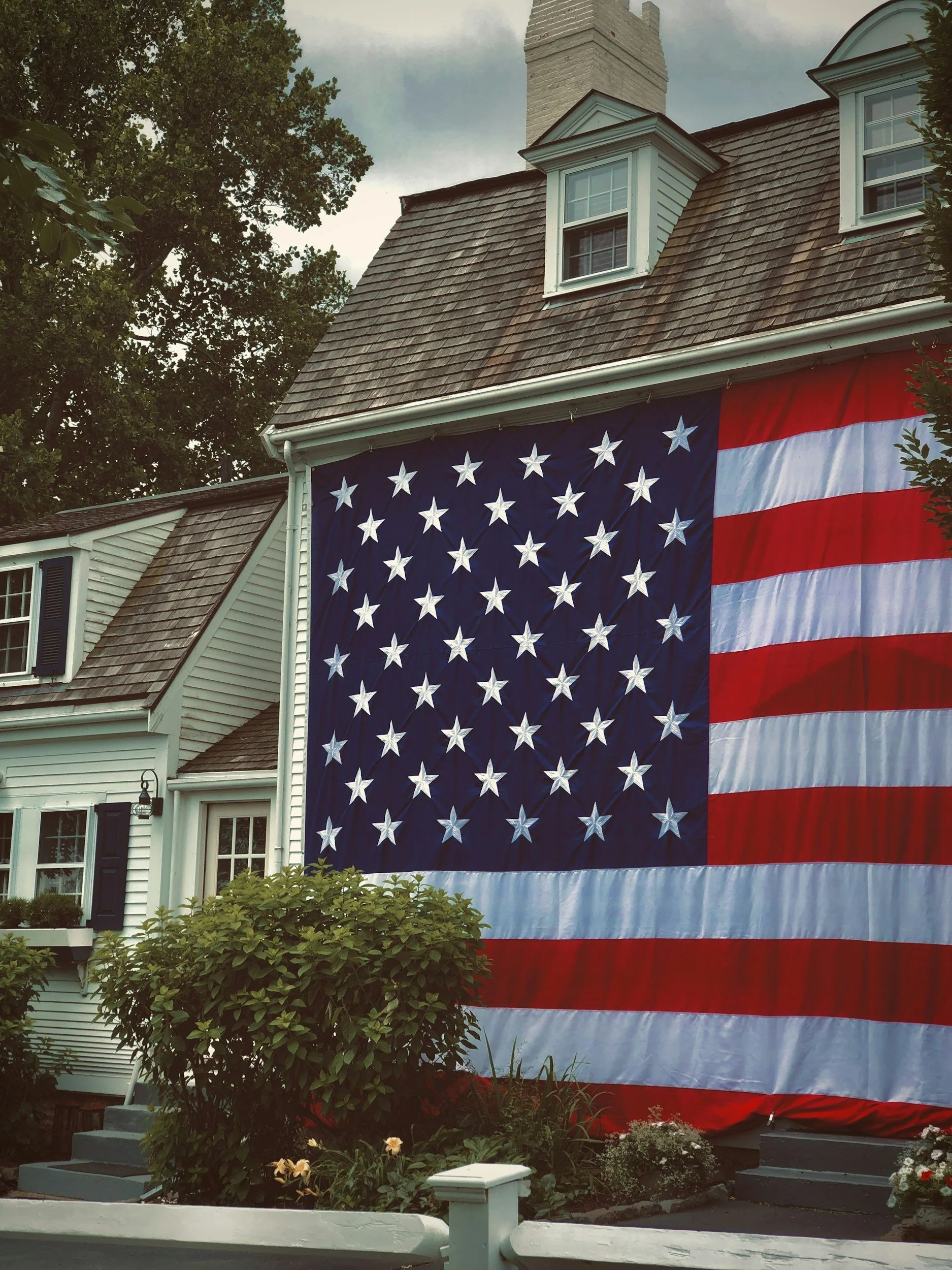 A large American flag hanging from the side of a house with white siding, dark shutters, and a pitched roof with dormer windows, surrounded by trees and a garden with bushes and flowers.