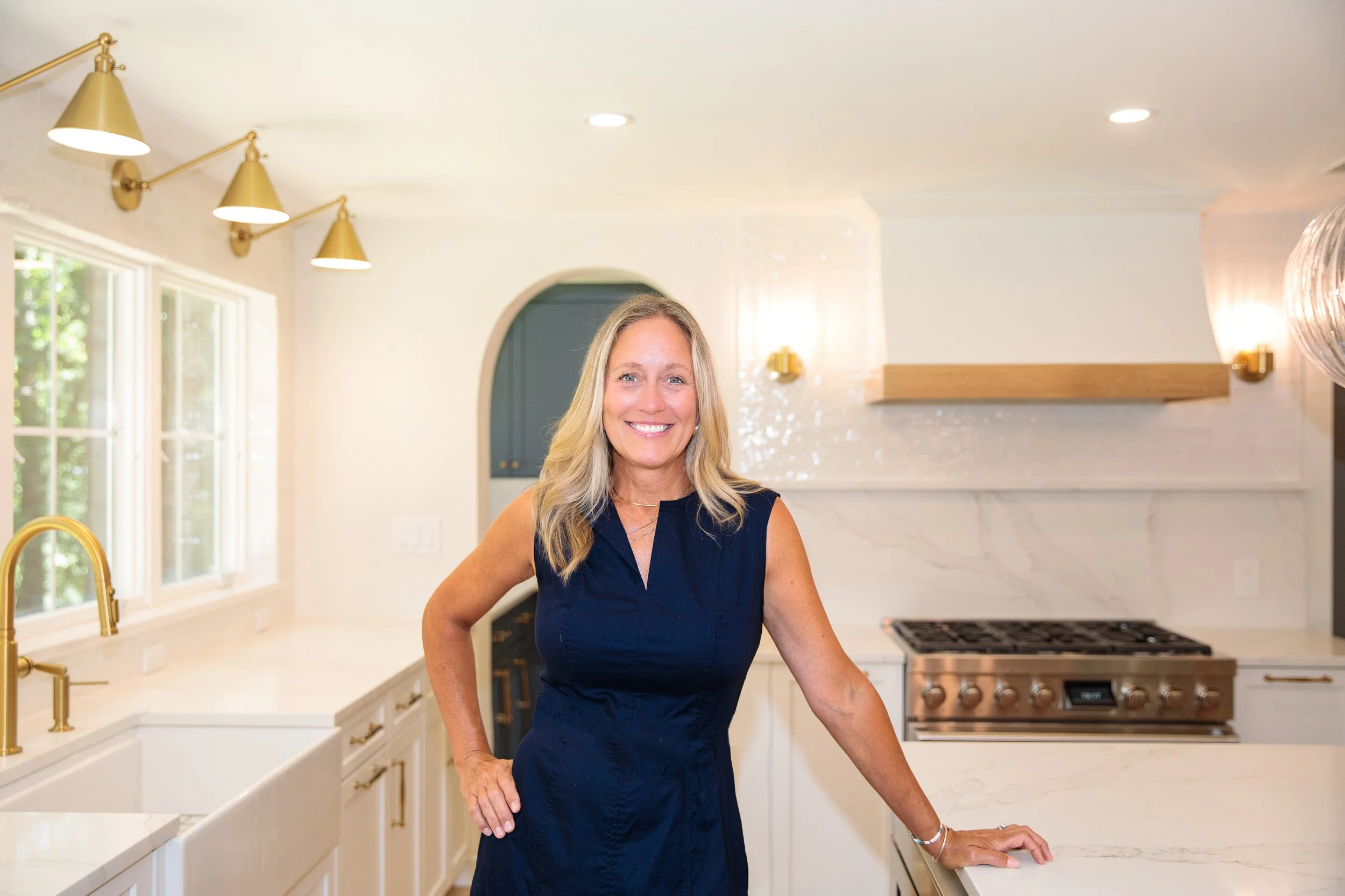 Woman in a navy blue dress smiling in a bright, modern kitchen with white cabinets, gold fixtures, and marble countertops.