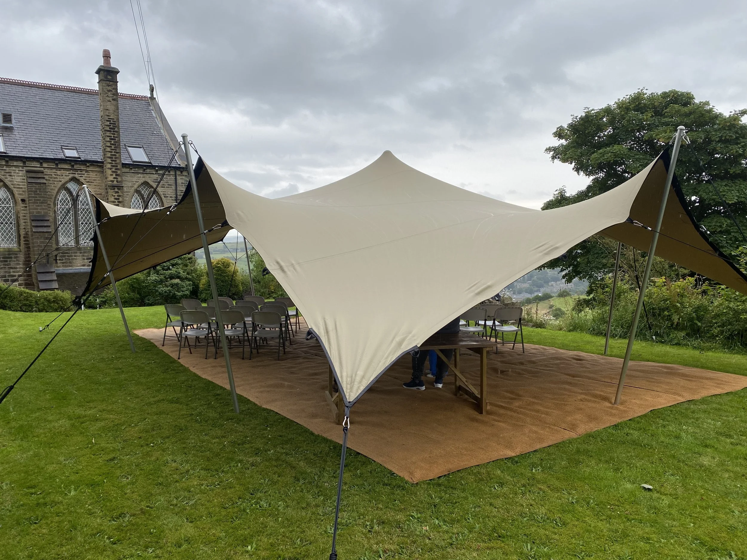 Outdoor event setup with a large beige canopy tent, tables, and chairs on a grassy lawn with a historic stone building and trees in the background.