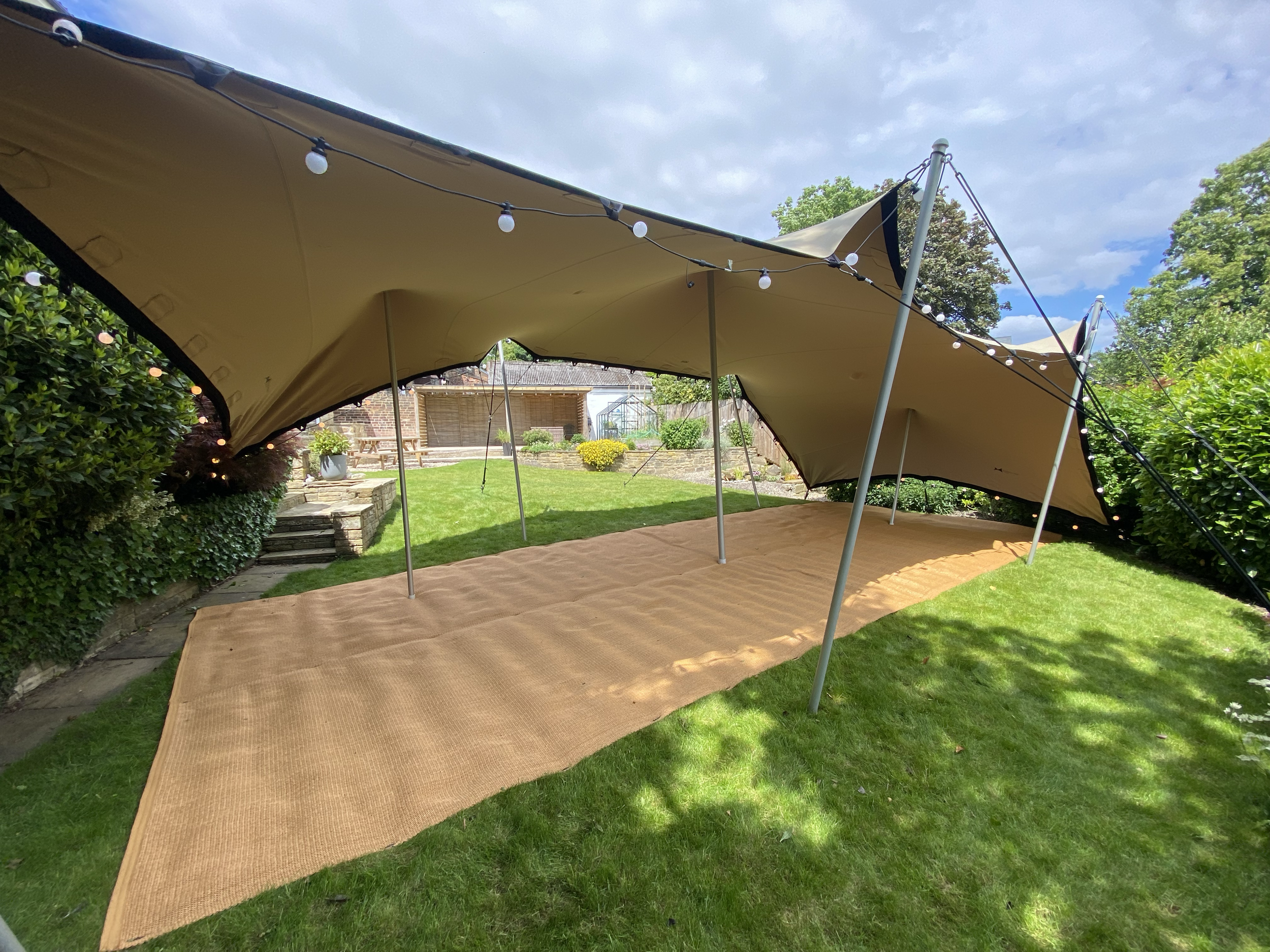 Large outdoor beige canopy tent set up on a lush green lawn in a backyard, with string lights hanging from it, surrounding bushes, and a small garden and patio area in the background.