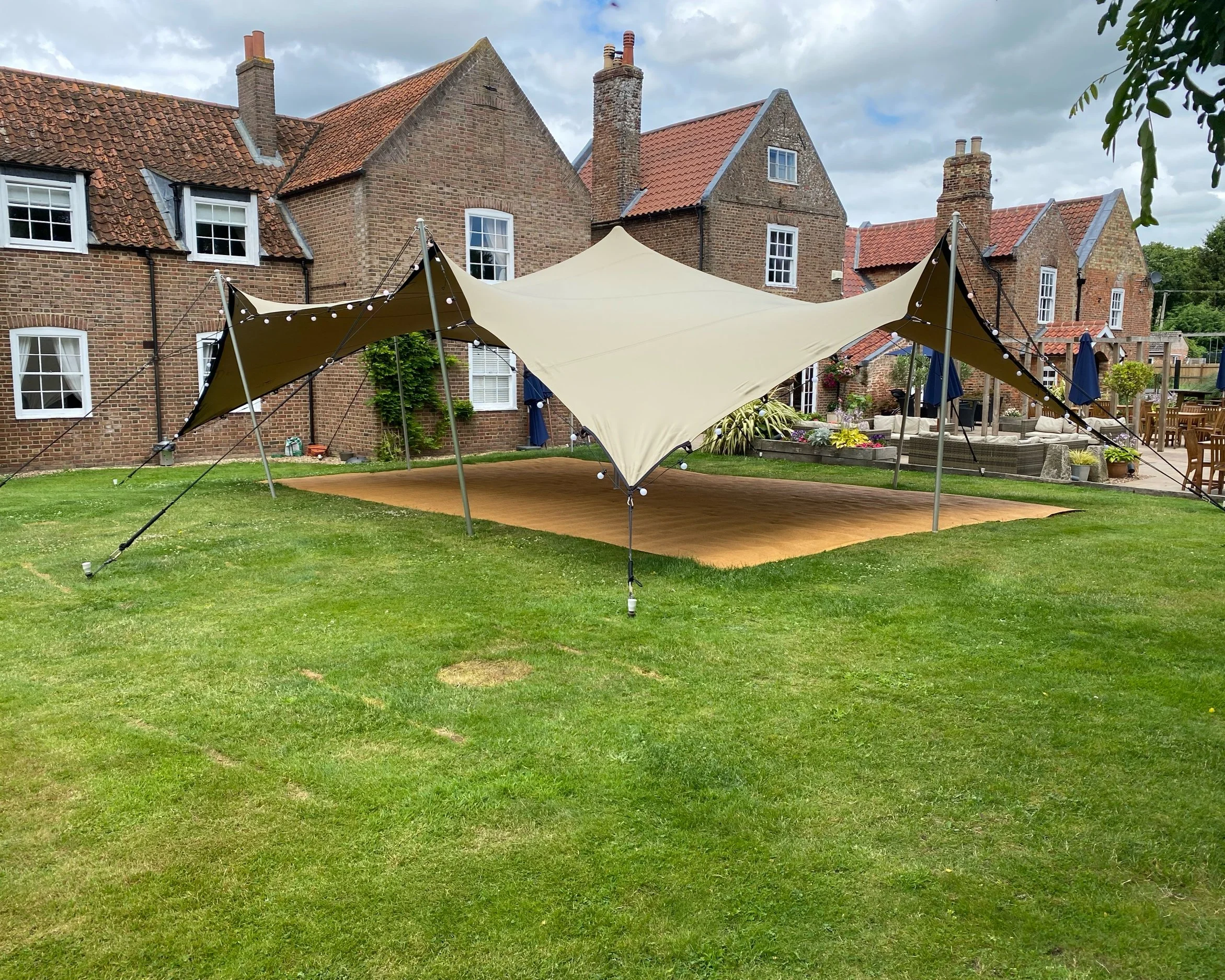 Large beige shade sail tent with string lights, set up over a wooden deck on a green lawn in a backyard with brick houses in the background.