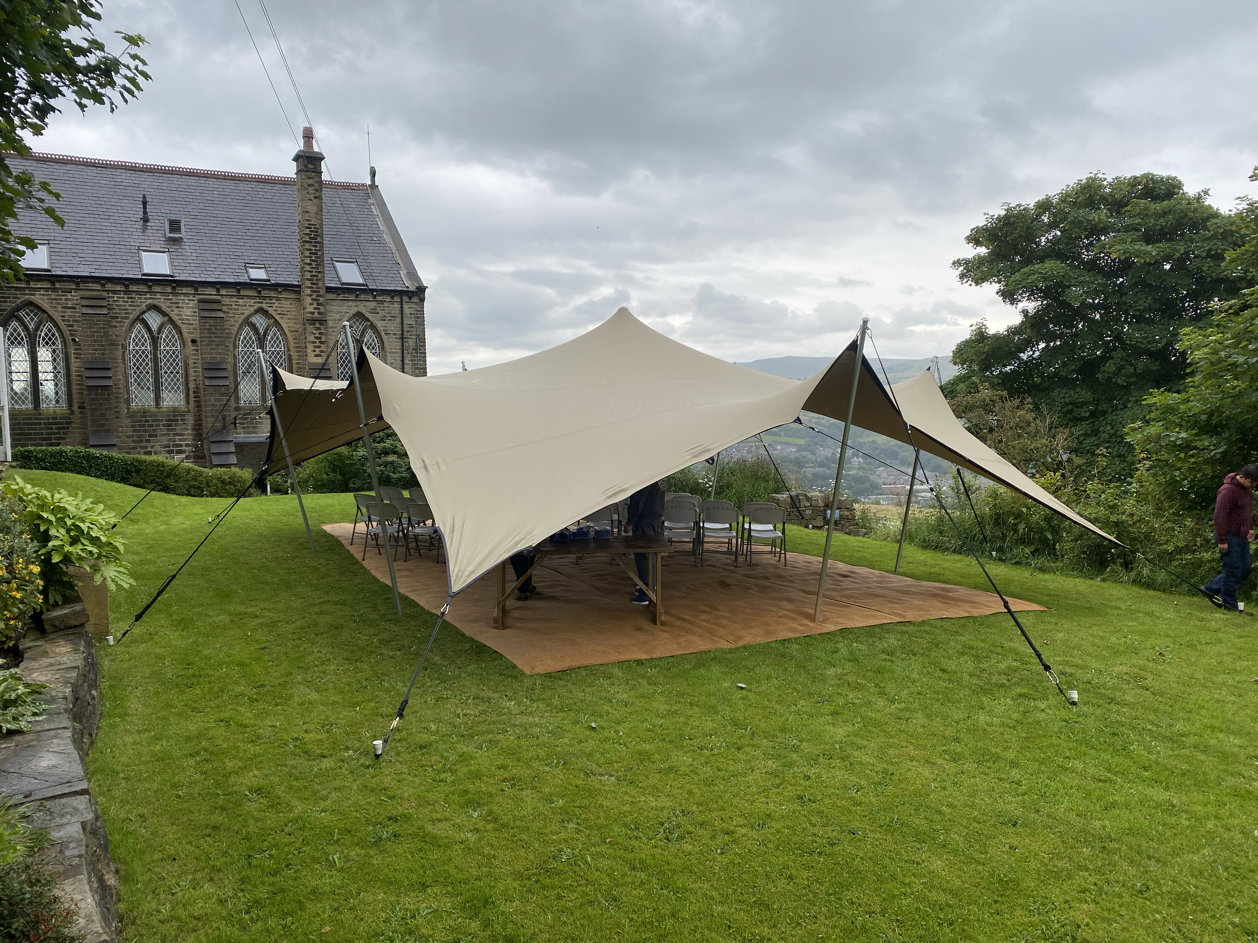Outdoor event setup with a large beige canopy tent on a grassy lawn near an old stone building, with chairs underneath and a brown floor covering, cloudy sky overhead.