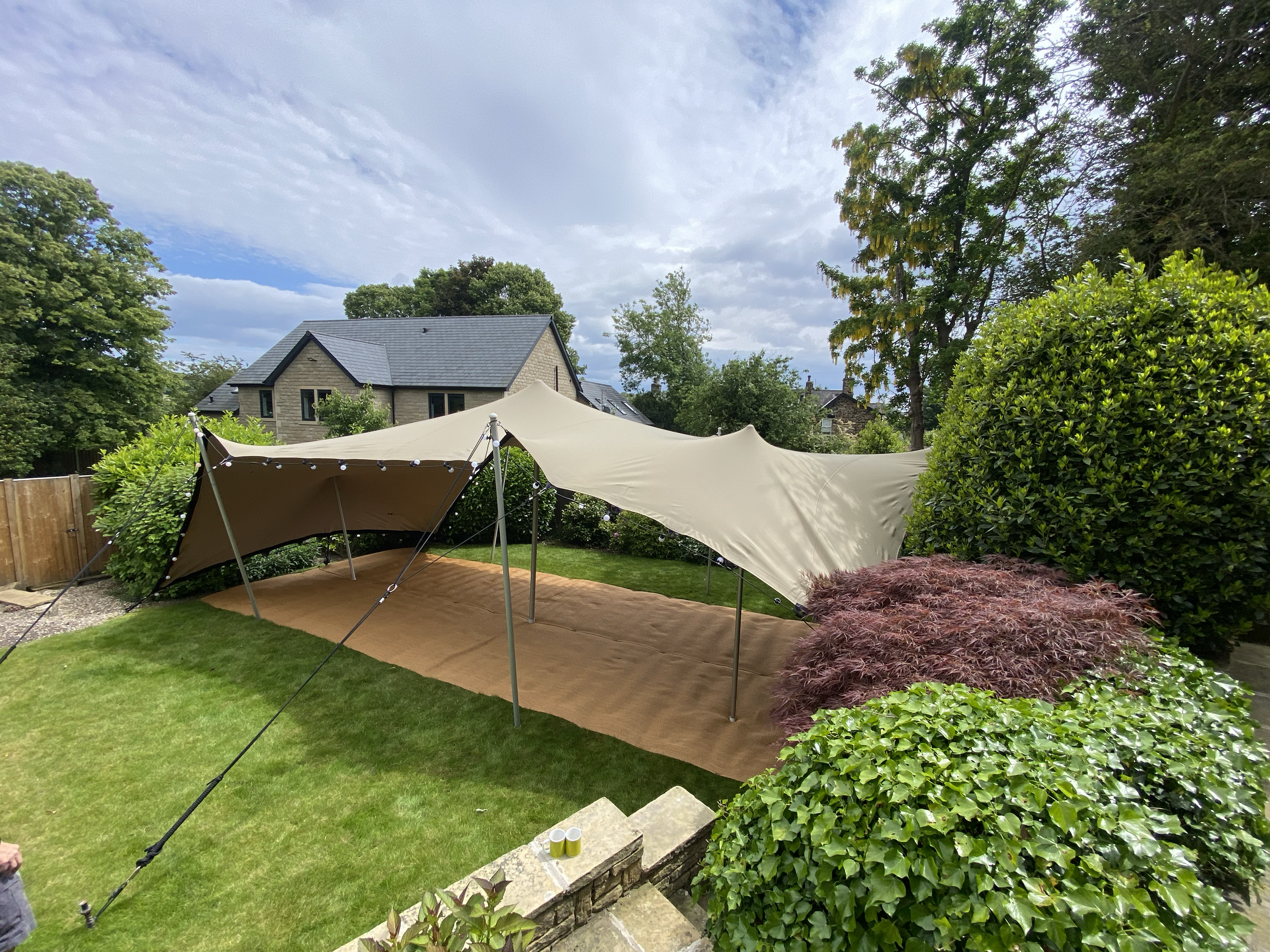 Outdoor backyard scene with a beige canopy tent set up on a brown outdoor rug, surrounded by green bushes and trees, with a house in the background and cloudy sky above.