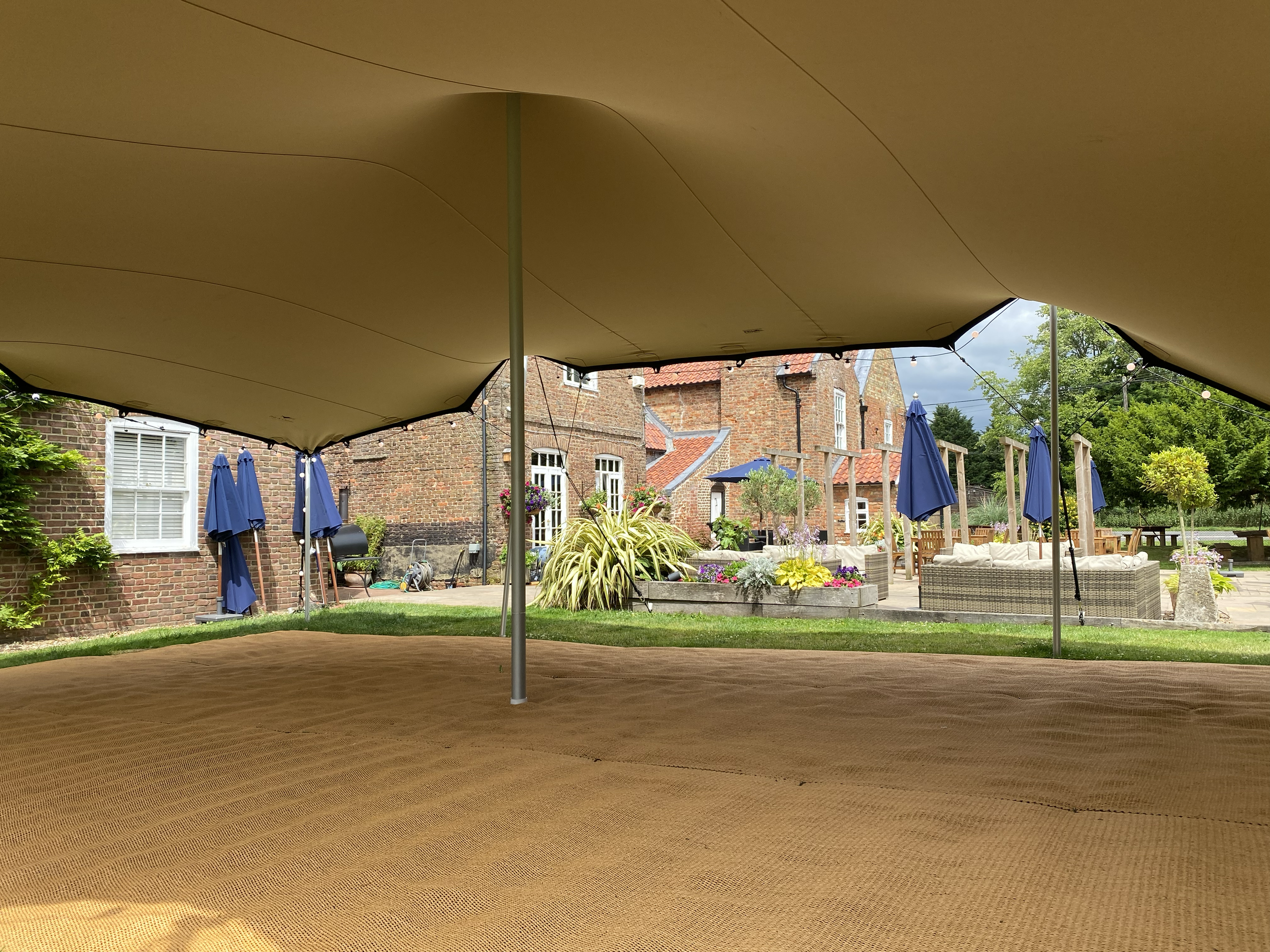 View from inside a beige canopy tent, looking out onto a well-maintained backyard with brick houses, blue patio umbrellas, outdoor seating, and lush greenery.