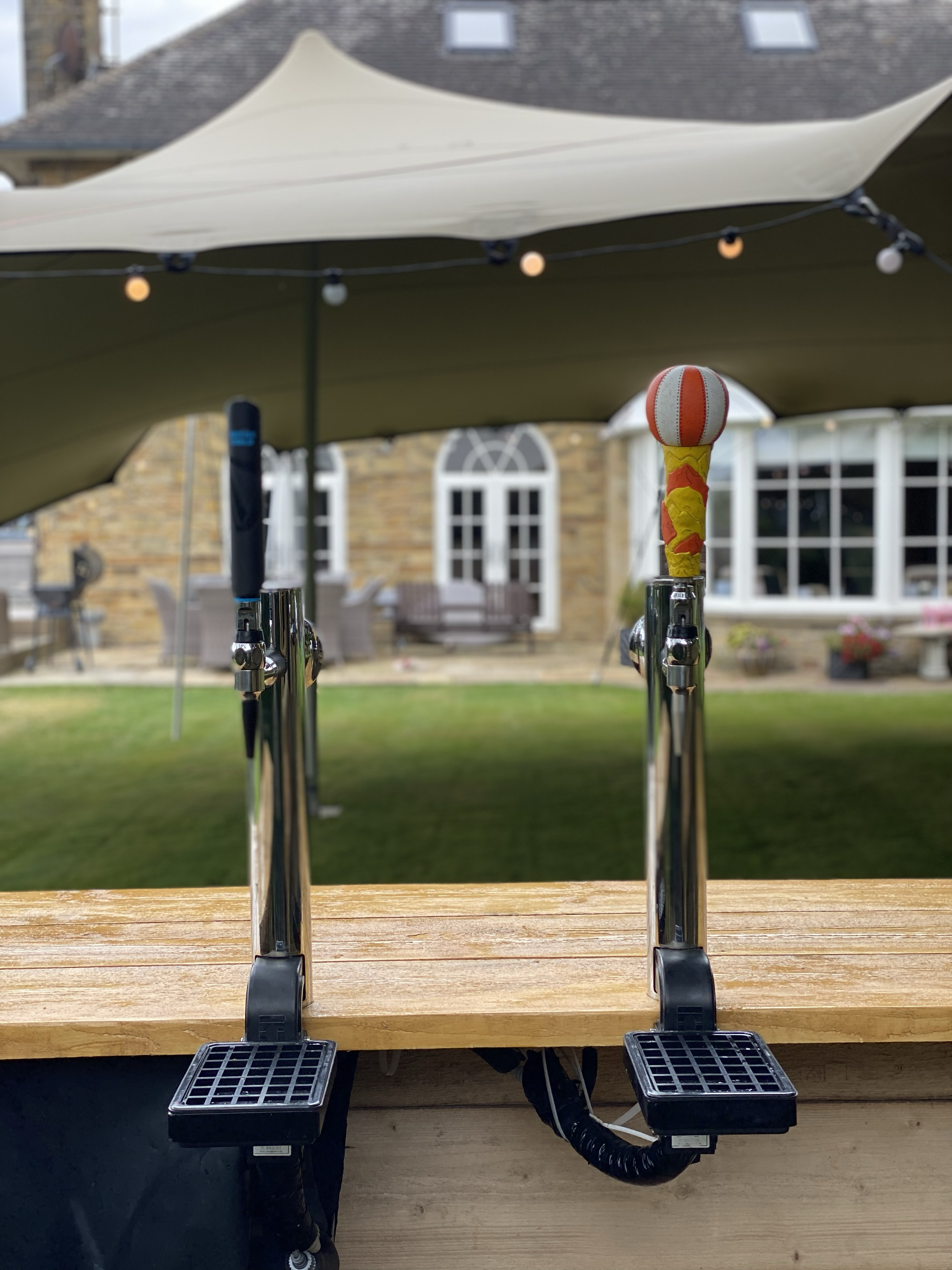 Two beer taps installed on a wooden bar, with a backyard patio and house in the background, shaded by a large canopy, and string lights overhead.