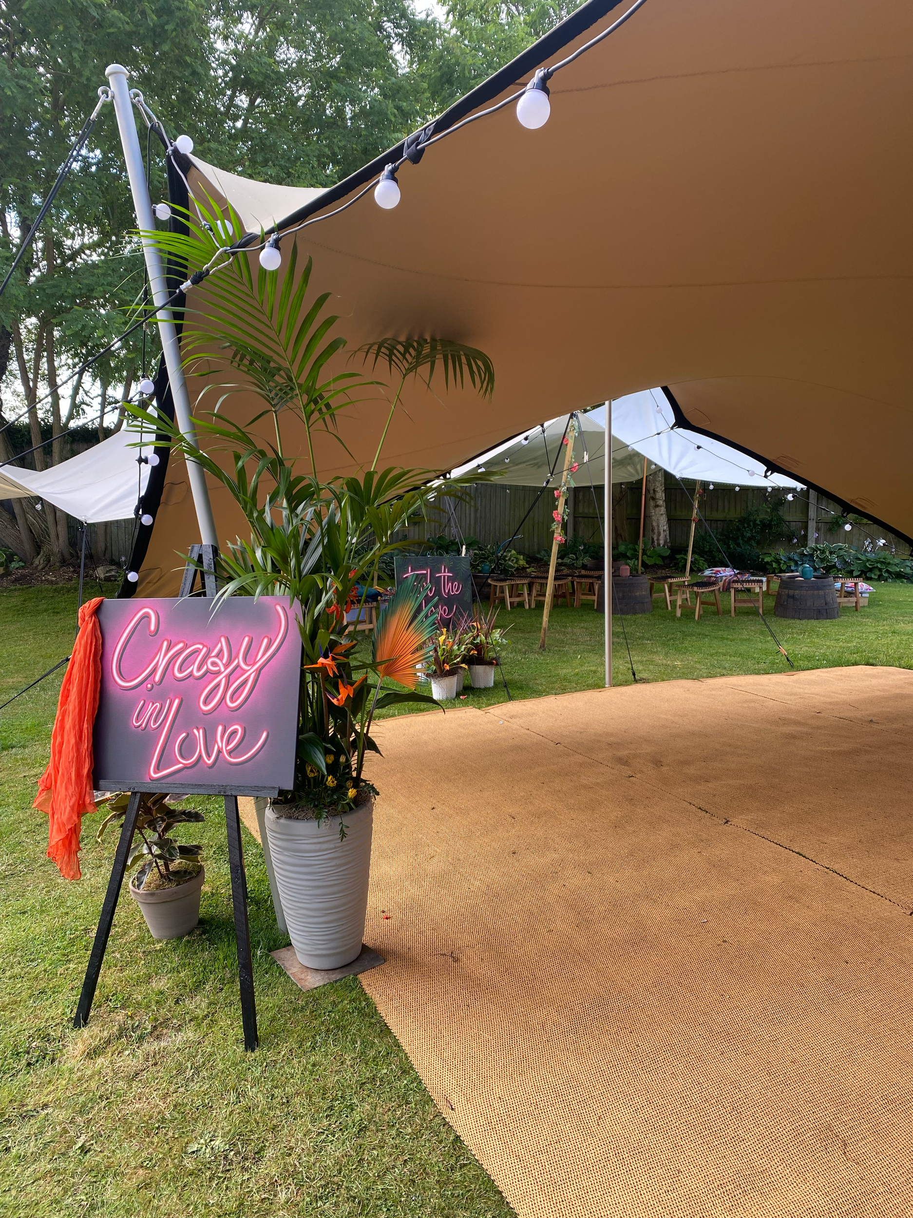 Outdoor scene with a beige canopy, string lights, potted plants, a pink neon sign that says "Creak in Love," and a sandy pathway.